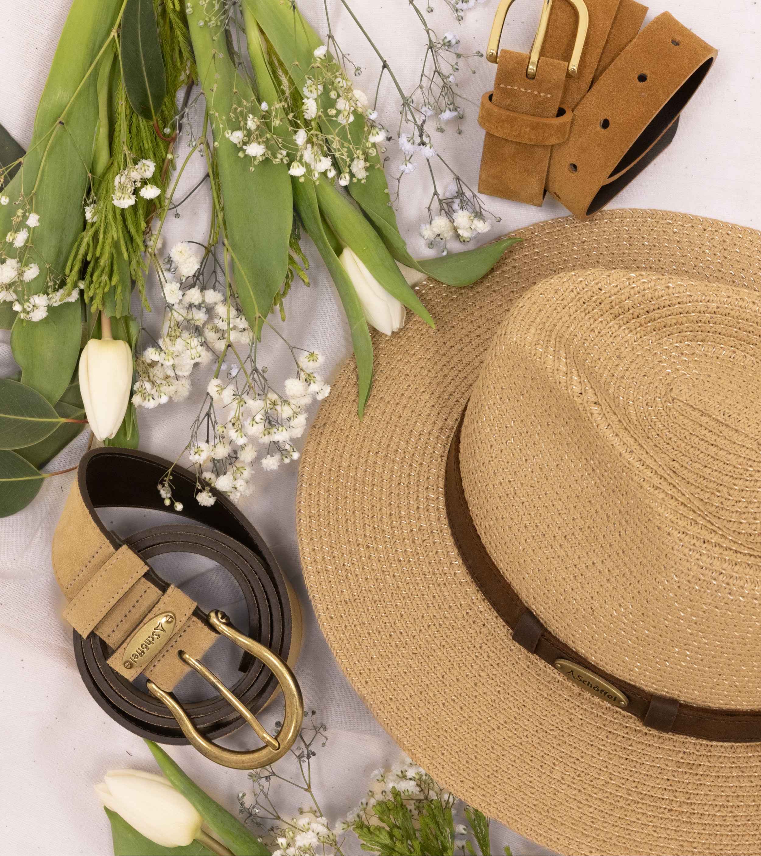 A flat lay image of belts and a straw hat with green and white foliage added as decoration.