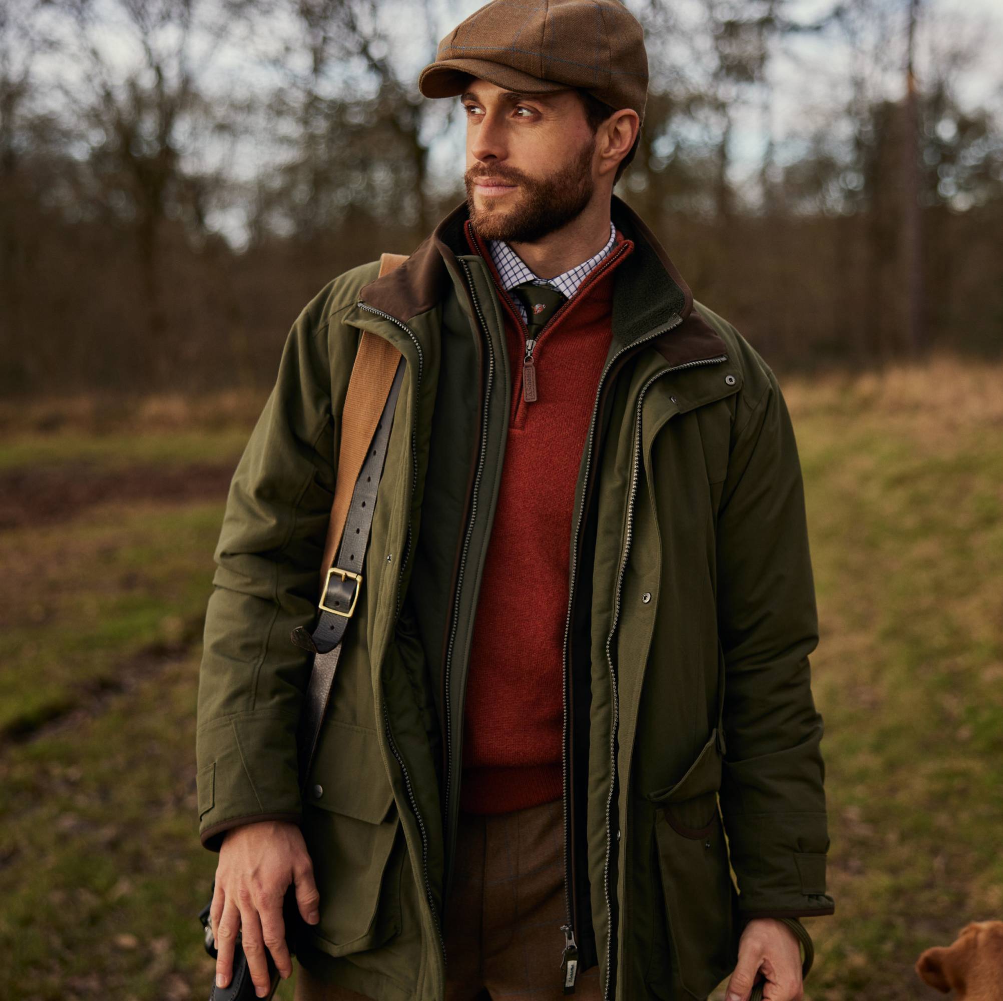 A man is seen looking to his left whilst wearing a green shooting jacket, and a red 1/4 zip knitwear. A check shirt can be seen underneath with a green tie.