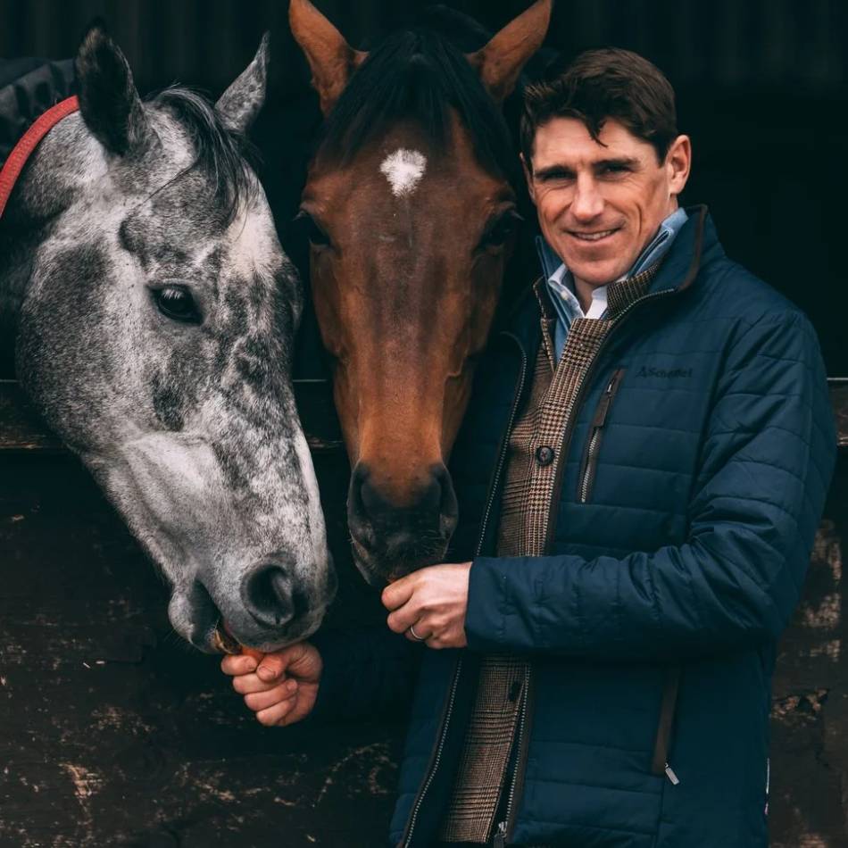 A man is looking forward and smiling whilst feeding a grey horse and a brown horse carrots. The man is wearing a navy padded jacket over a tweed check waistcoat.