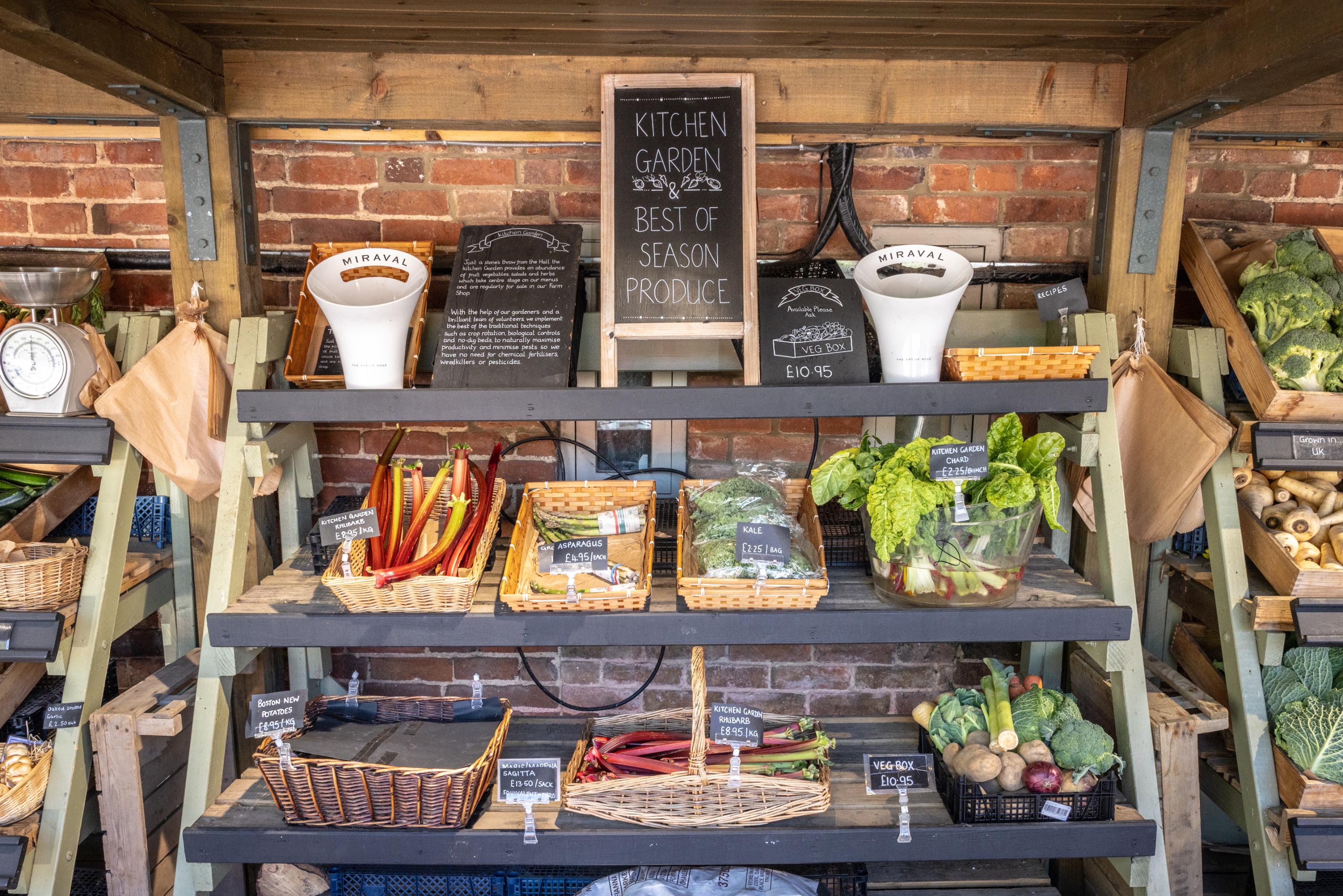 A close-up of a stall featuring kitchen garden seasonal produce. There are woven baskets of rhubarb, asparagus, spinach, and mixed vegetables.