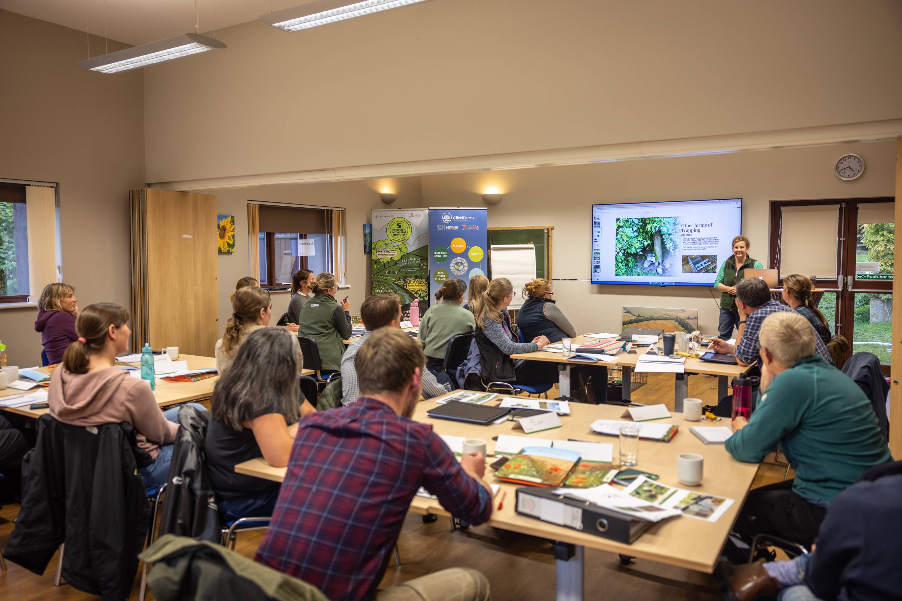 Image shows a classroom of people at large desks. They are all looking at a screen at the front of the classroom.