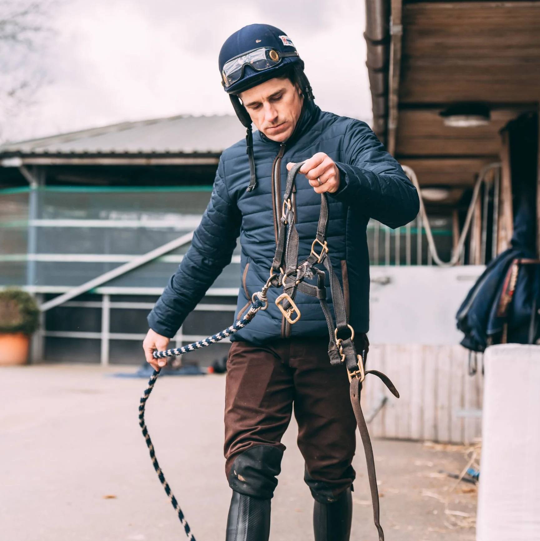 A man is holding a horses head collar and lead rain in a stable courtyard. The man wears a navy padded jacket, brown breeches and a navy helmet and cover.