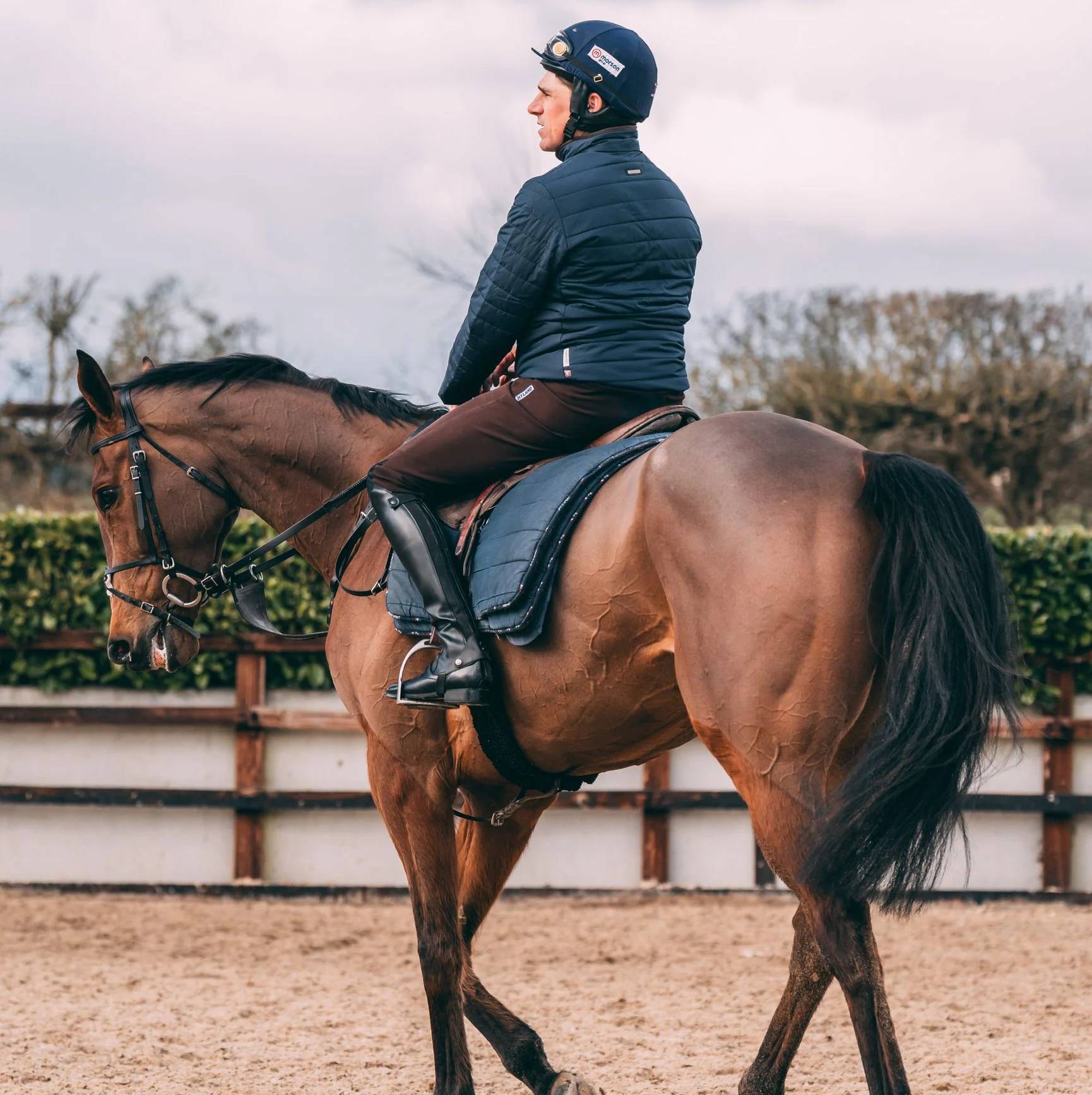 A side view of a man riding a brown horse in a sanded paddock.