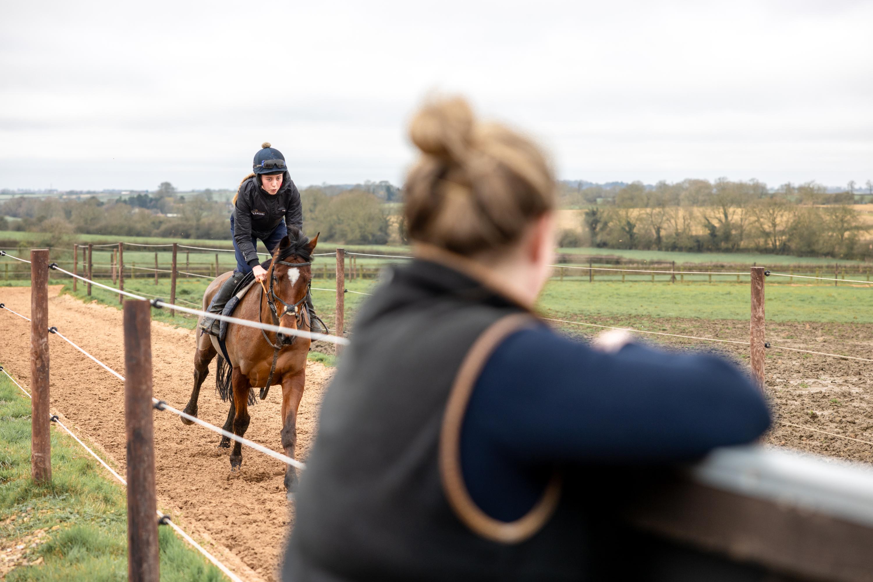 A female is seen riding a brown horse along a sandy track in the gallop position. The rider wears a riding hat with a navy cover and in the foreground a watcher looks at the riding coming forward in a gallop.