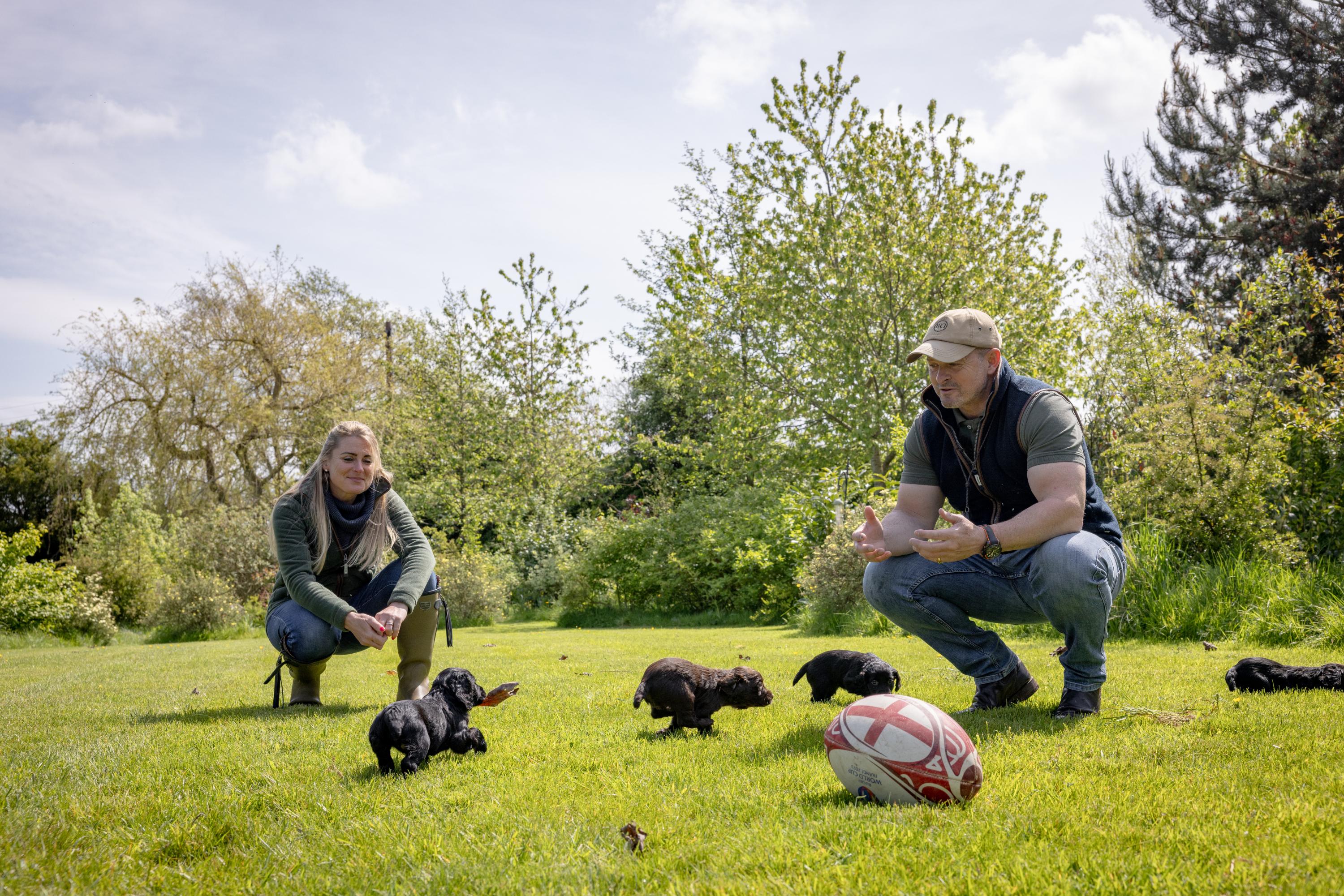 A man and a woman are both seen crouching down whilst interacting with puppies as they play and run around a grass park or field.