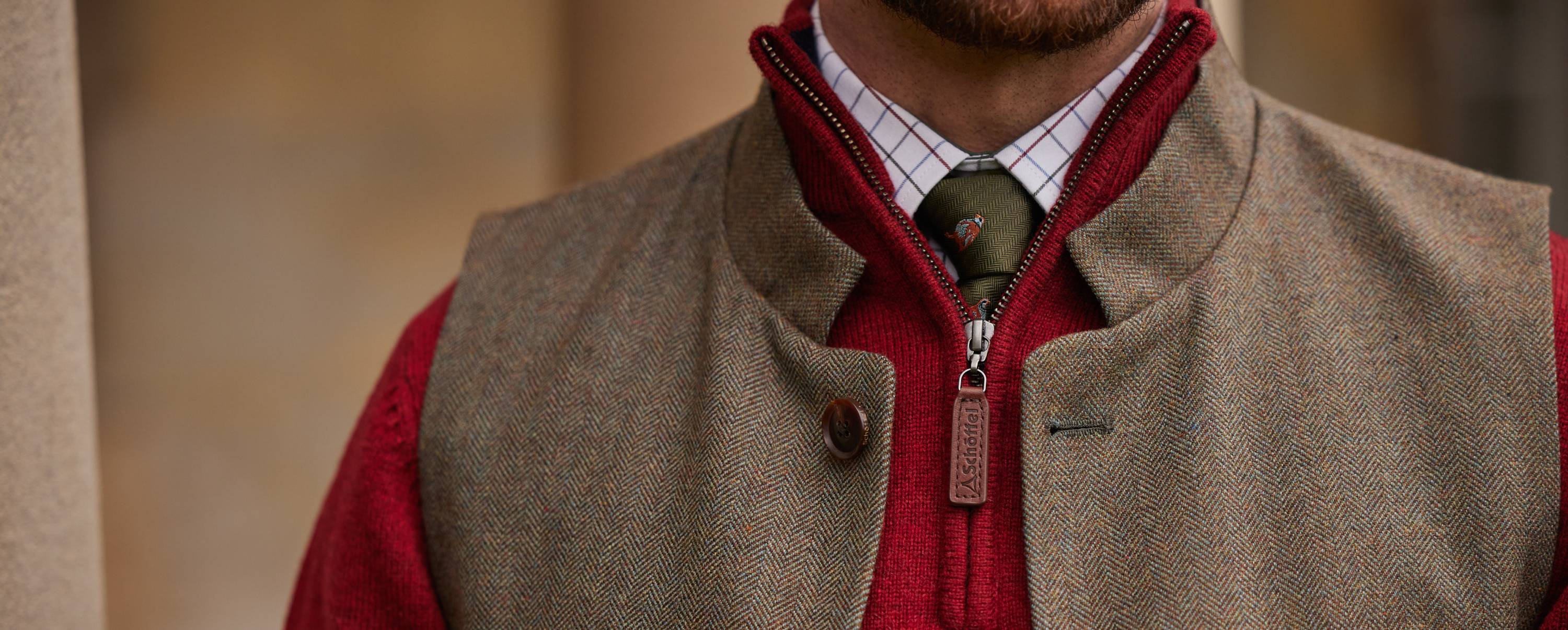 A close-up of a mans collar area. The man wears a tweed waistcoat, red 1/4 zip knitwear, check shirt and a green tie.