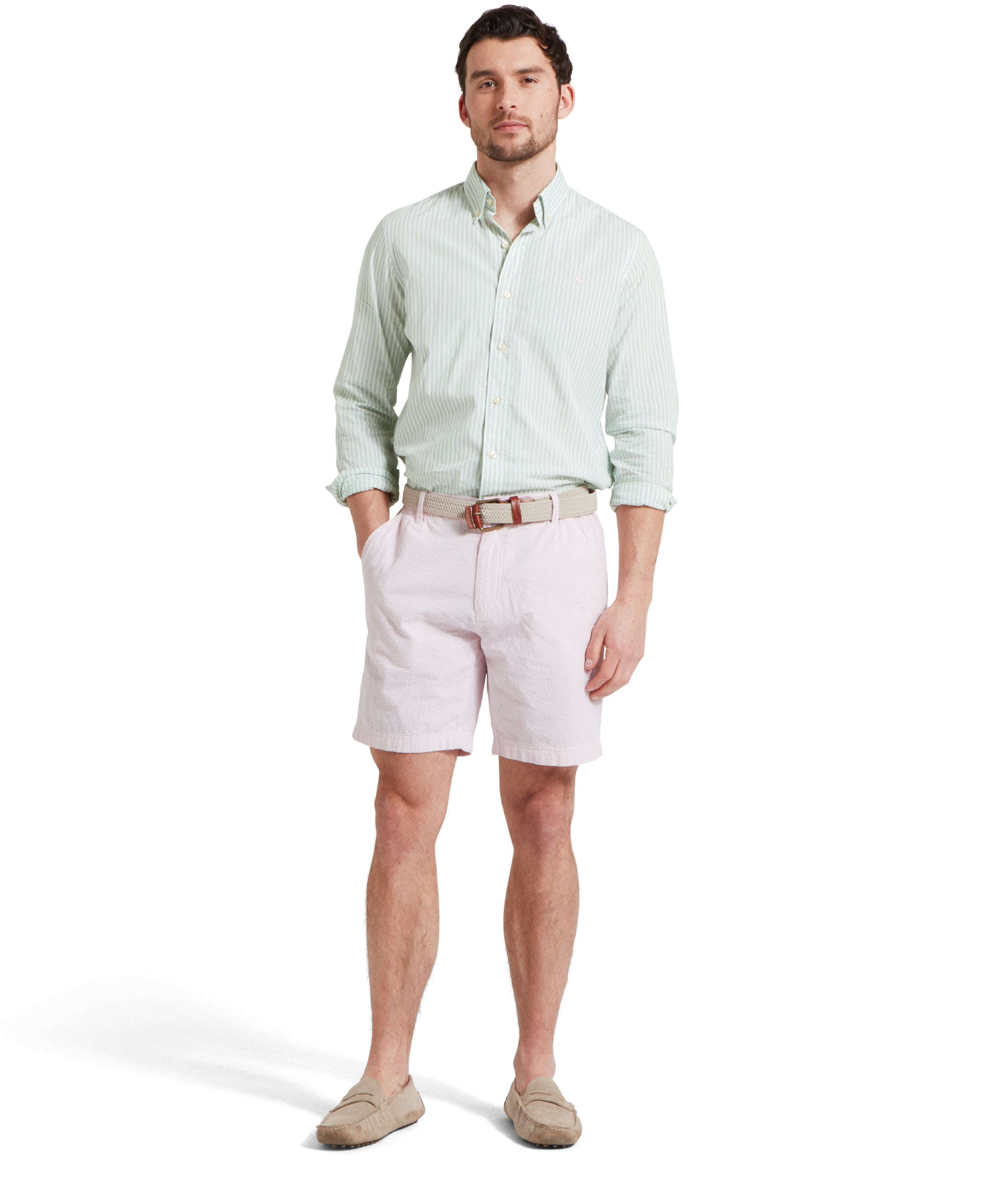 A full height image of a male model wearing a light pink shorts with a tucked in light green and white striped shirt. It is paired with a beige woven belt and tan loafers. He is standing in a relaxed pose with one hand in his pocket.