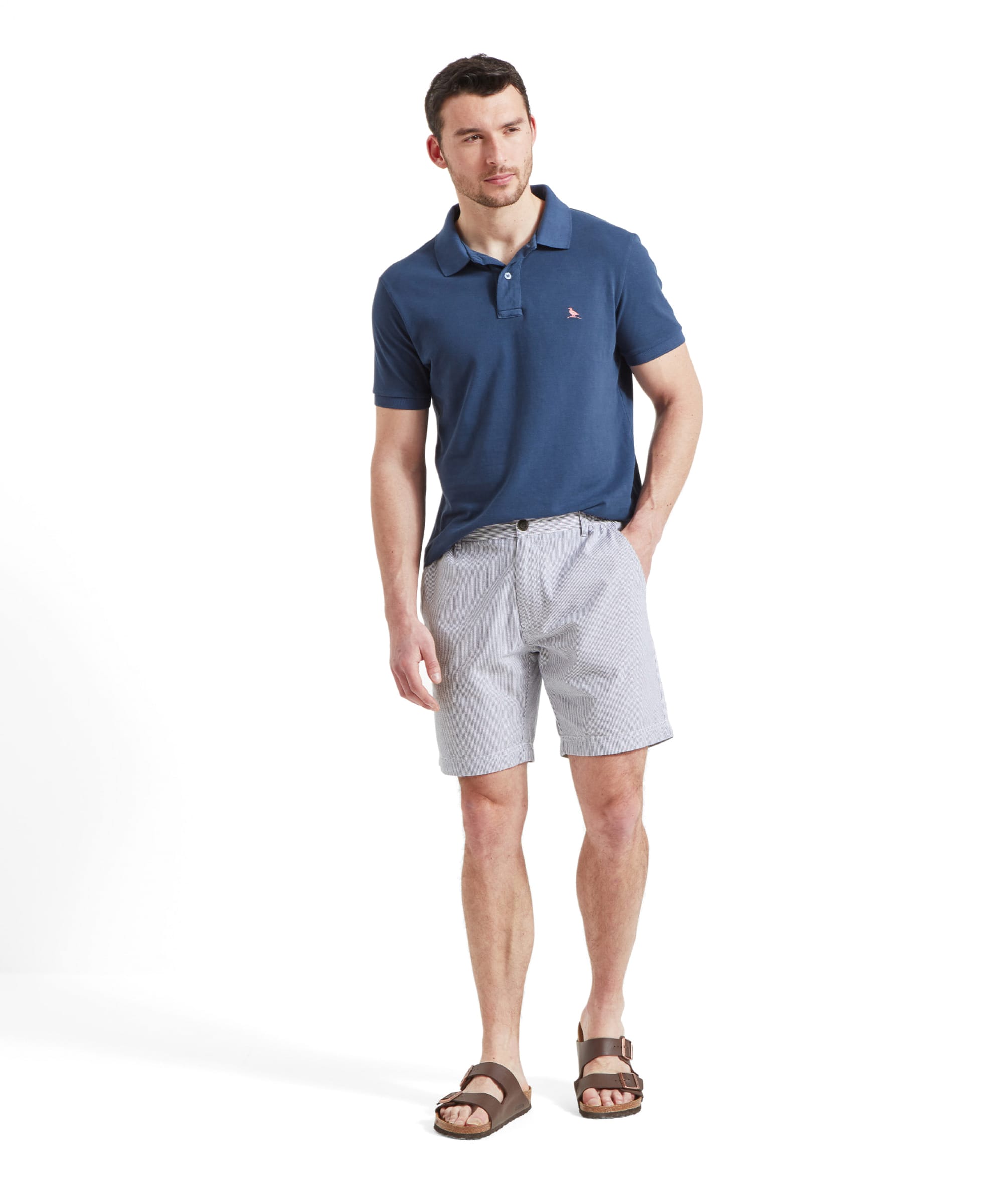 A full-body shot of a man wearing a blue polo shirt tucked into the Schöffel Abersoch Shorts for Men in Navy, with his hands in his pockets, standing casually and looking to the side.