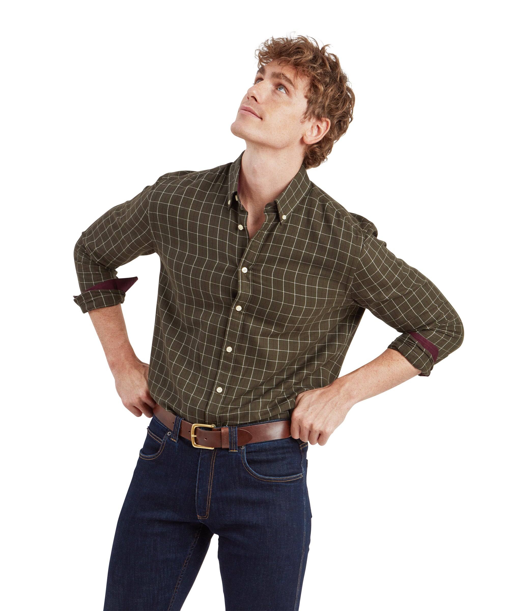 A man wearing a Schöffel Aldeburgh Tailored Shirt for Men in Green with the sleeves rolled up to the elbows. He has curly brown hair and is looking upwards with his hands placed on his hips. The shirt features a subtle grid pattern, and he pairs it with dark blue denim jeans and a brown leather belt.