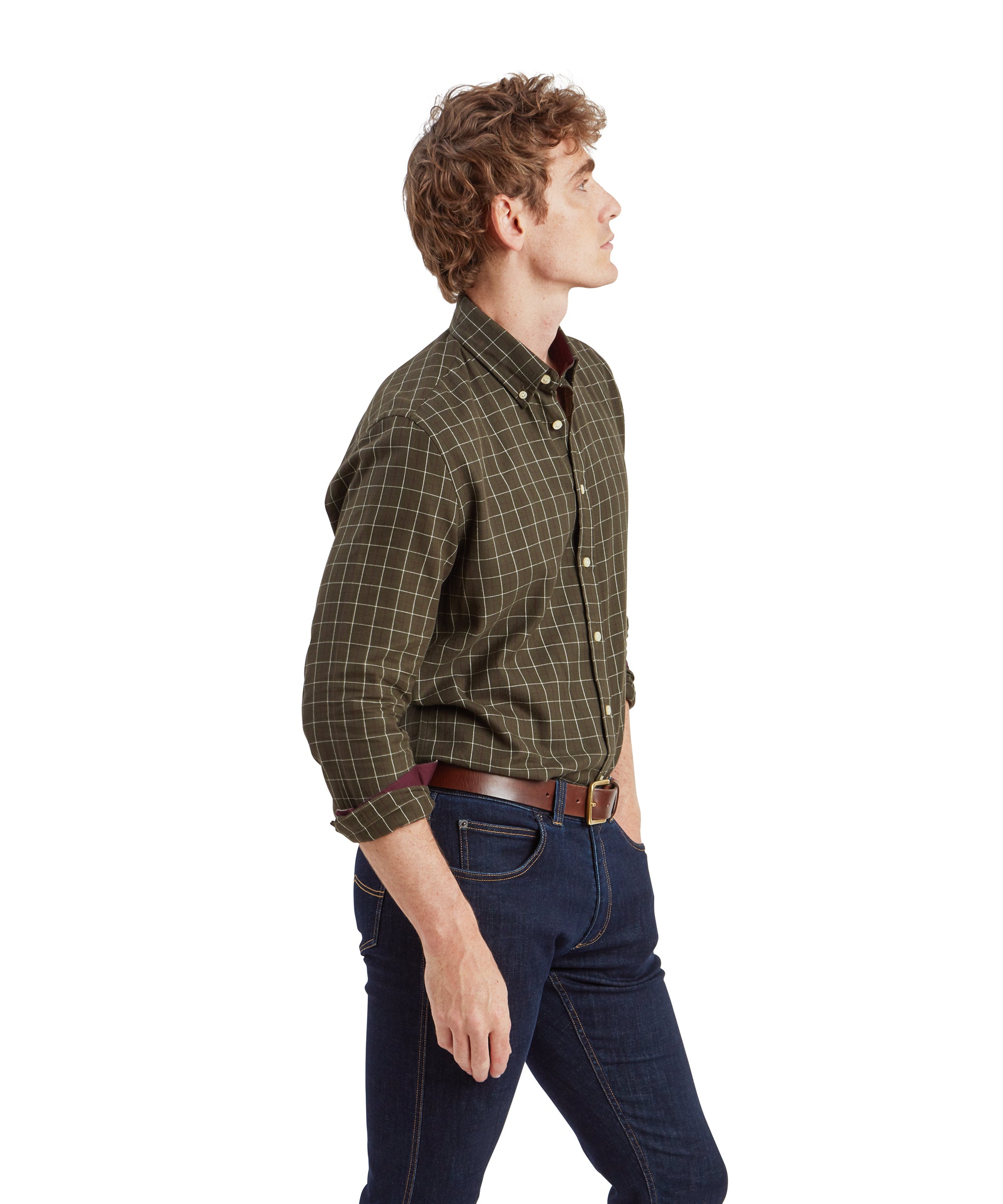 A side profile of the man in the Schöffel Aldeburgh Tailored Shirt for Men in Green. He is looking slightly upward with his hands in the pockets of his dark blue jeans. The shirt’s checkered pattern is clearly visible, and the burgundy inner cuff contrasts against the green.