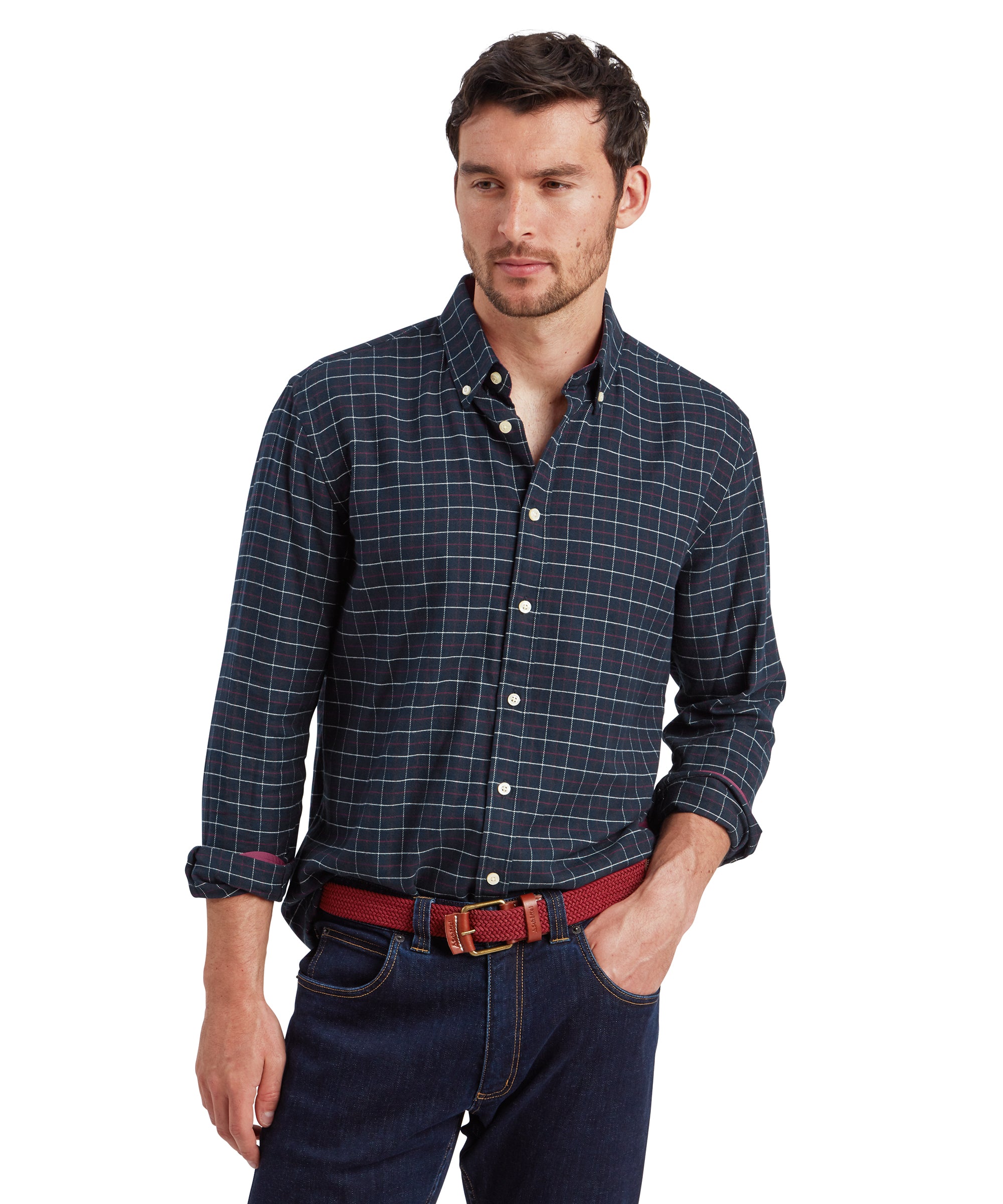 A male model is wearing a Schöffel Aldeburgh Tailored Shirt for Men in Navy with long sleeves. The shirt has a classic fit and a button-down collar. The model has his left hand in his jean pocket and is wearing a red woven belt with gold buckle detailing. He stands facing the camera with a neutral expression.