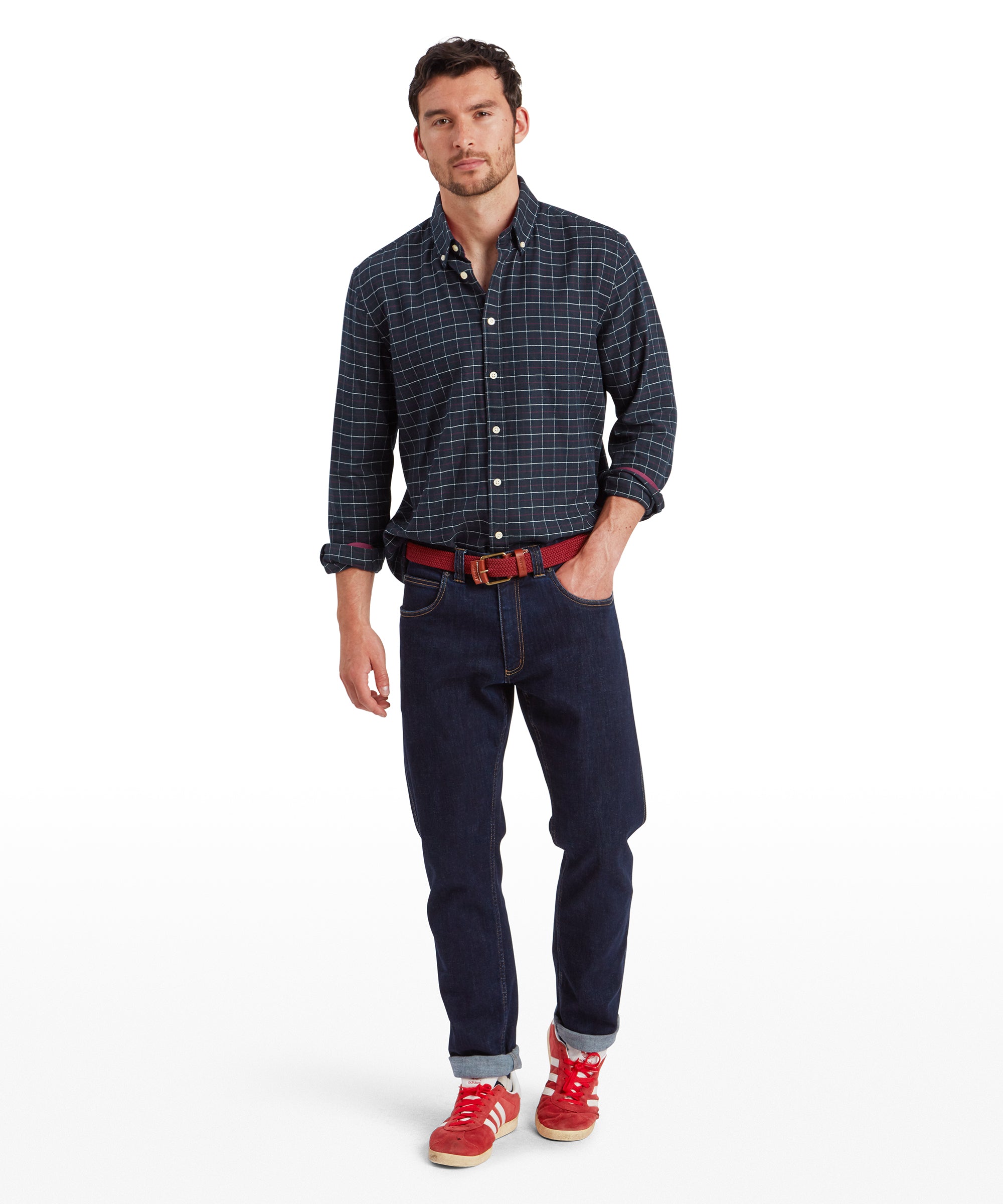 A full-body shot of the male model wearing the same Schöffel Aldeburgh Tailored Shirt for Men in Navy, paired with dark blue jeans and red trainers. He has his left hand in his pocket and is looking slightly down and to his right. His casual stance and red woven belt complement the overall outfit.