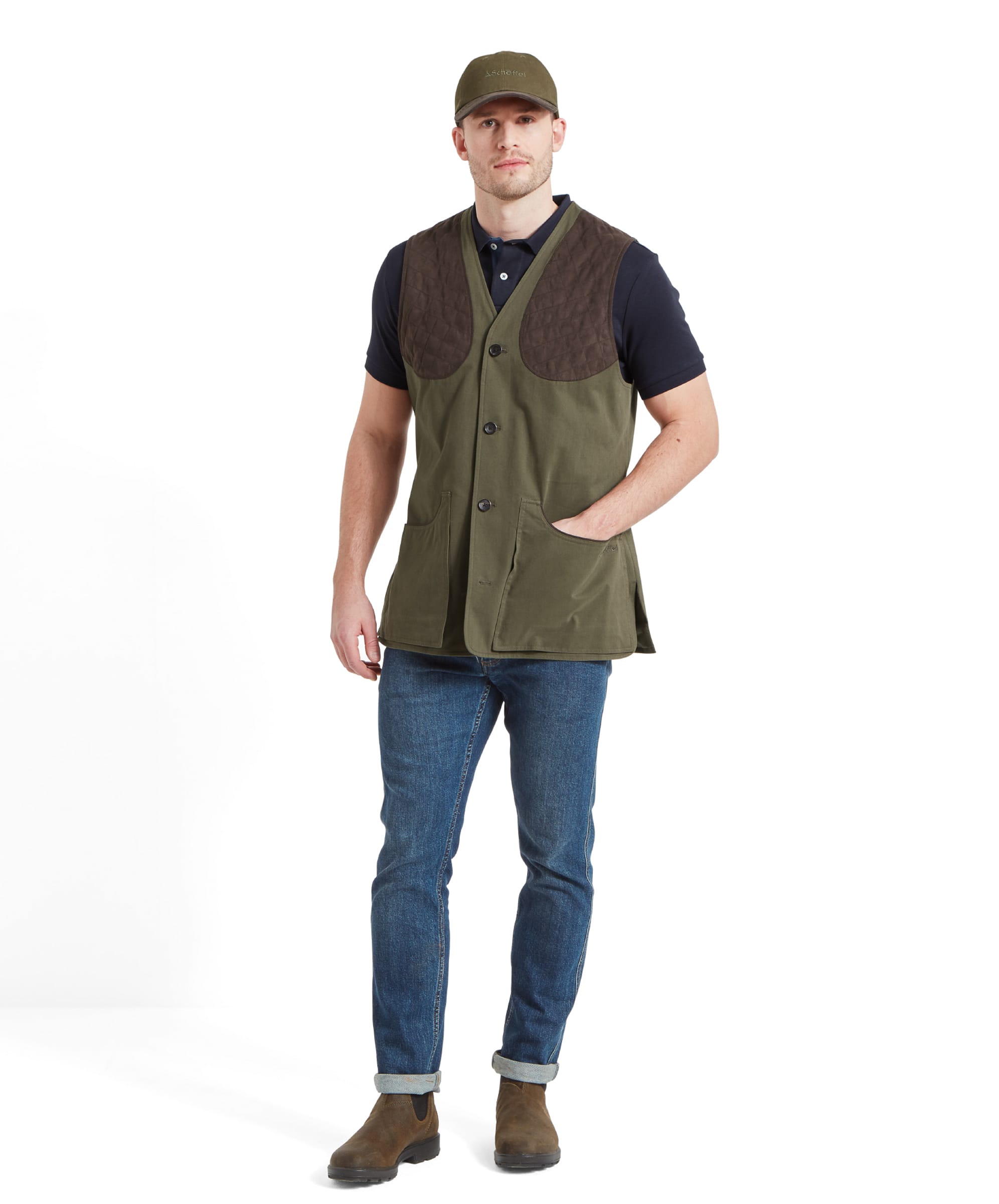 A full-body shot of a man wearing a Schöffel All Season Shooting Vest for Men in Green with quilted shoulder pads over a navy blue shirt, blue jeans, and brown boots. He is standing casually with one hand in his vest pocket.