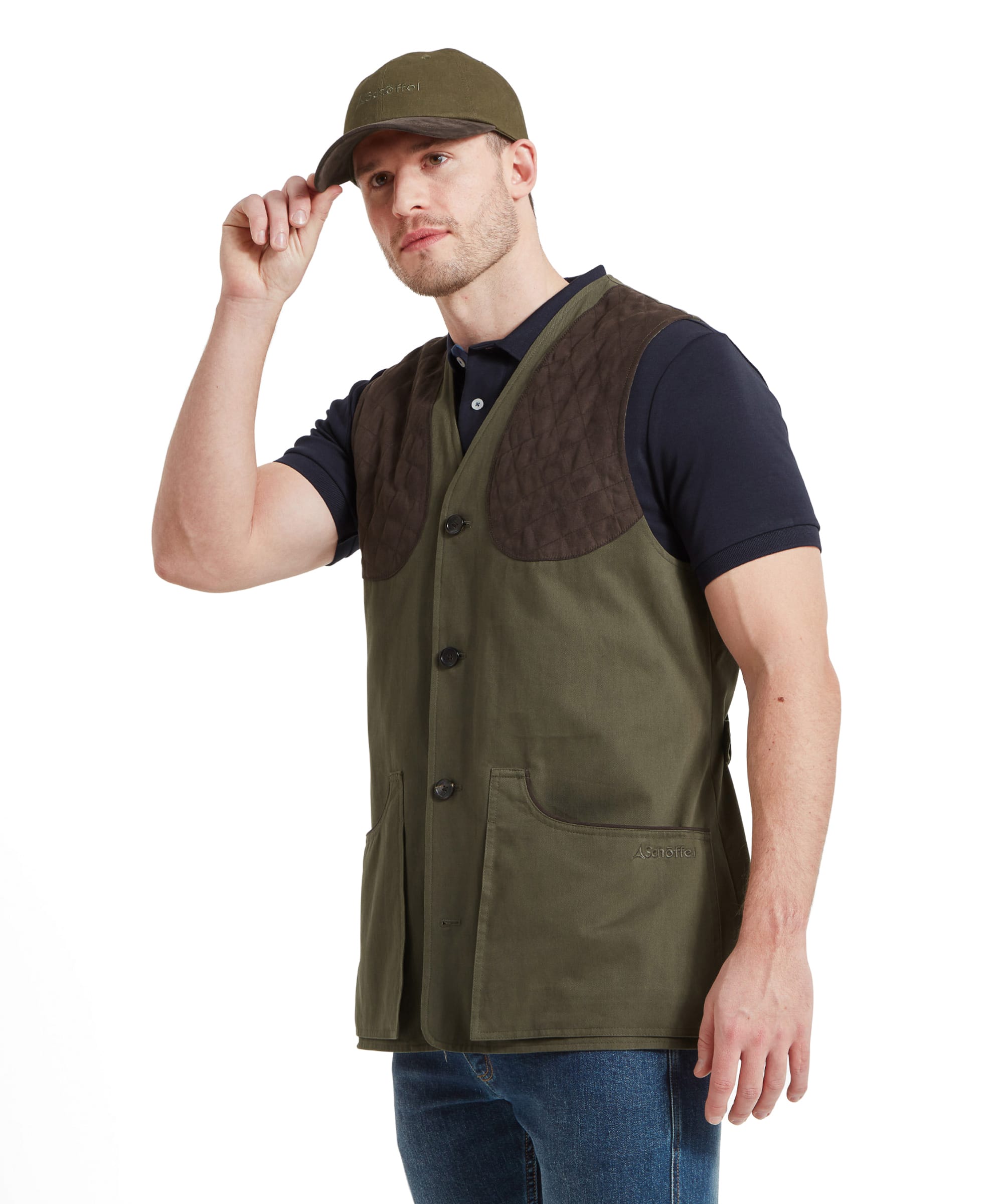 A man wearing a Schöffel All Season Shooting Vest for Men in Green with quilted shoulder pads, a navy blue shirt, and a green cap. He is adjusting his cap with one hand and has the other hand in his vest pocket.