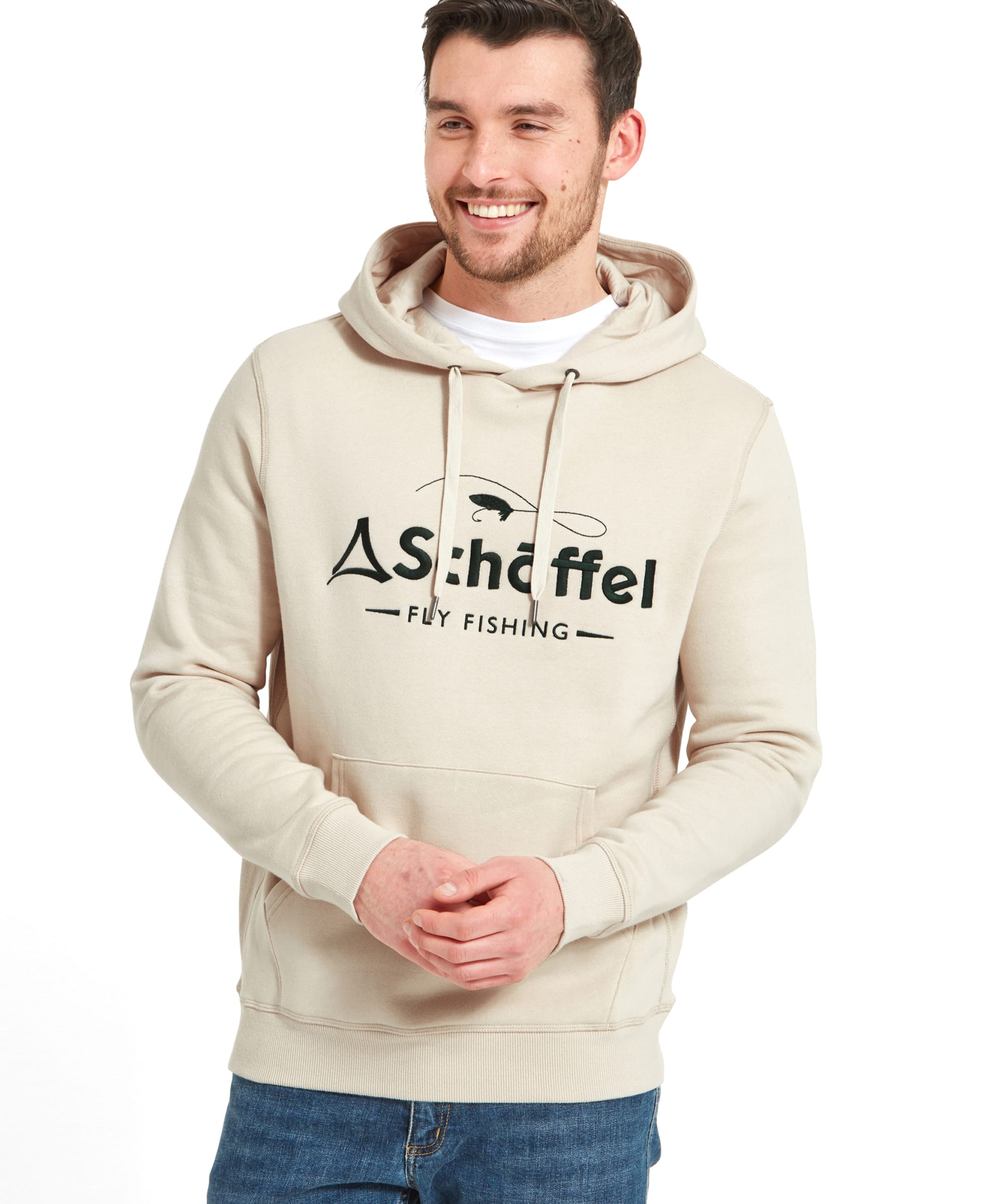 A man wearing a Schöffel Alness Hoodie for Men in Light Brown with the "Schöffel Fly Fishing" logo on the front, smiling and looking to the side. He is also wearing blue jeans.