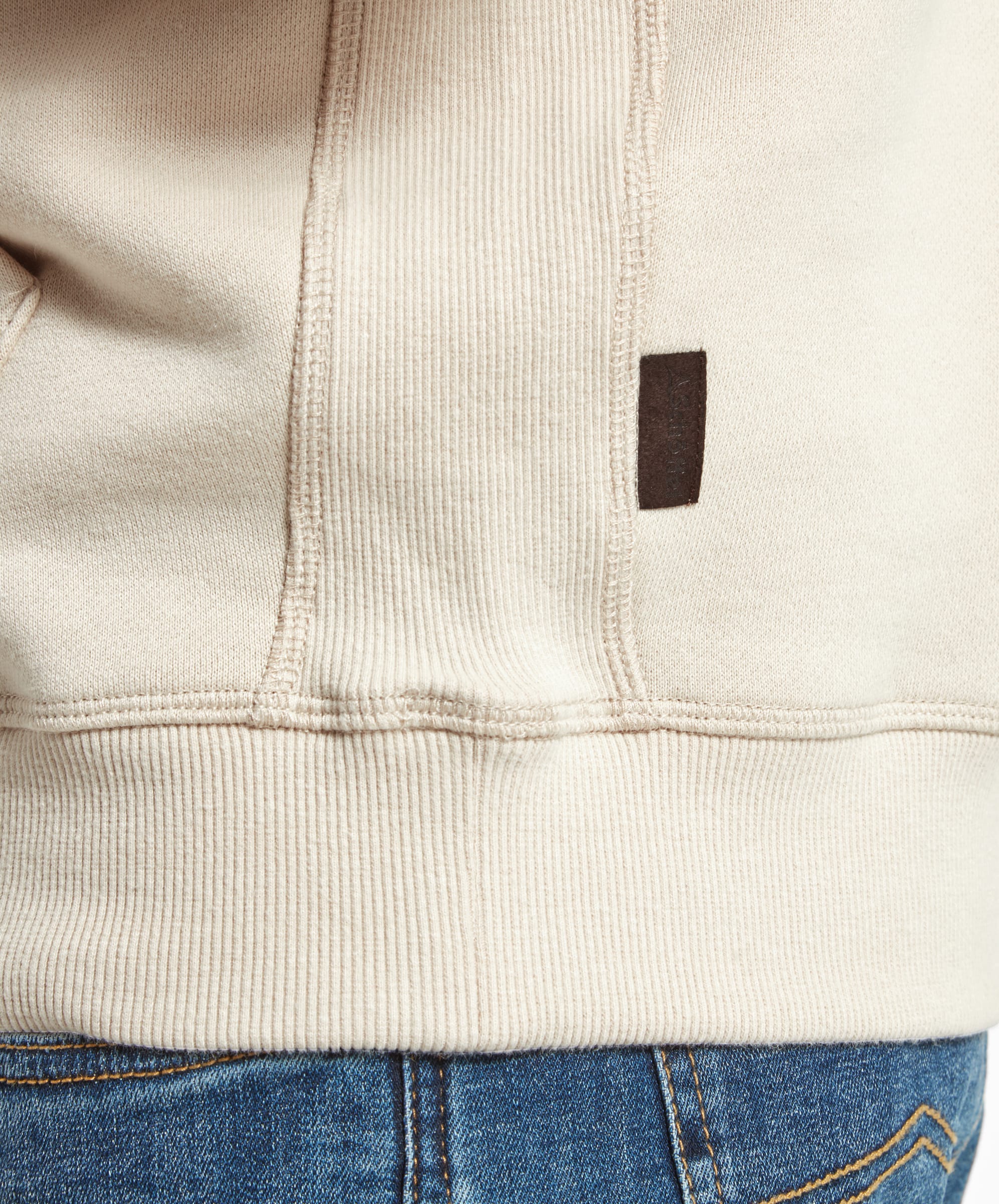 A close-up shot of the side seam detail of a Schöffel Alness Hoodie for Men in Light Brown, showing the stitching and a small dark brown fabric tag near the bottom hem.