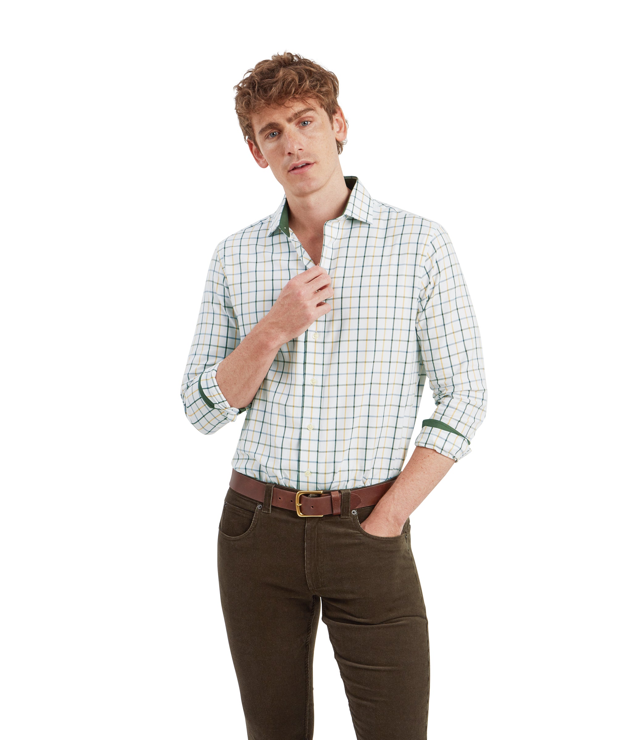 A young man with curly brown hair wearing a Schöffel Baconsthorpe Tailored Shirt for Men in Green with a checkered pattern in shades of green, white, and yellow. The shirt is tucked into olive-green corduroy pants with a brown leather belt. He is standing with his right hand partially tucked into his pocket, while his left hand adjusts the collar of the shirt. The man gazes slightly upward to his left with a thoughtful expression.