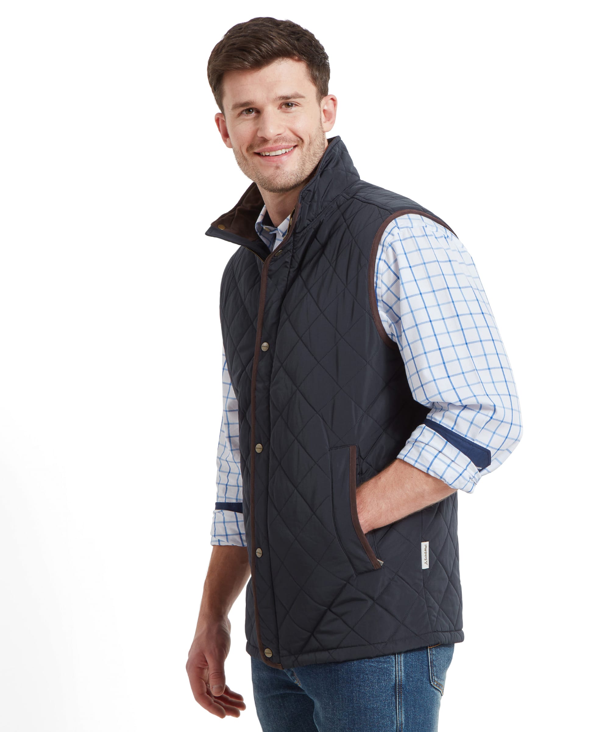 A side view of a man wearing a Schöffel Barrowden Quilt Gilet for Men in Blue with brown trim over a light blue and white checkered shirt, smiling and looking to the side with his hands in his pockets.