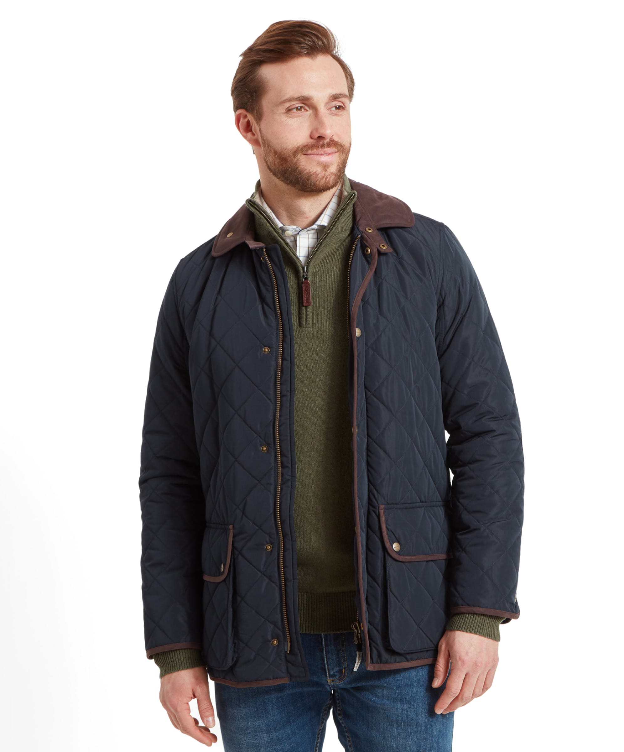 A man modeling a Schöffel Barrowden Quilt Jacket for Men in Blue, looking to the side with a slight smile. He wears a green sweater and blue jeans.