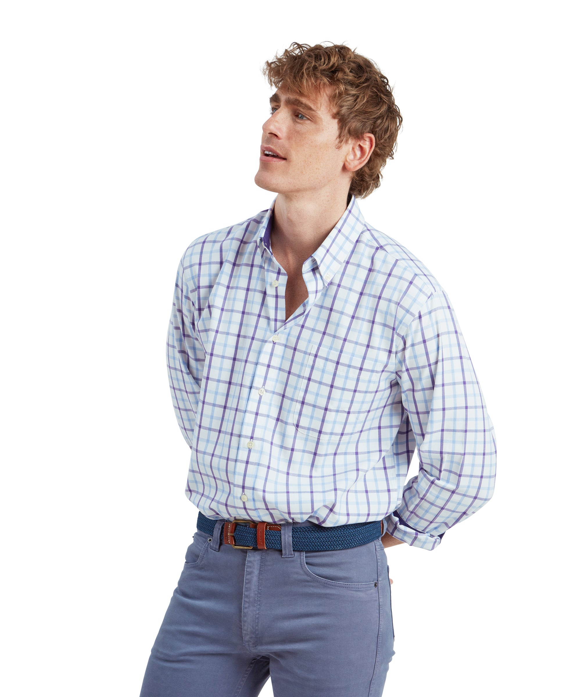 A side profile shot of the young man in the Schöffel Brancaster Classic Shirt for Men in Purple. He stands with his hands behind his back, looking slightly upwards with a relaxed expression. The shirt is neatly tucked into the slim-fit light blue pants, with the blue woven belt visible.