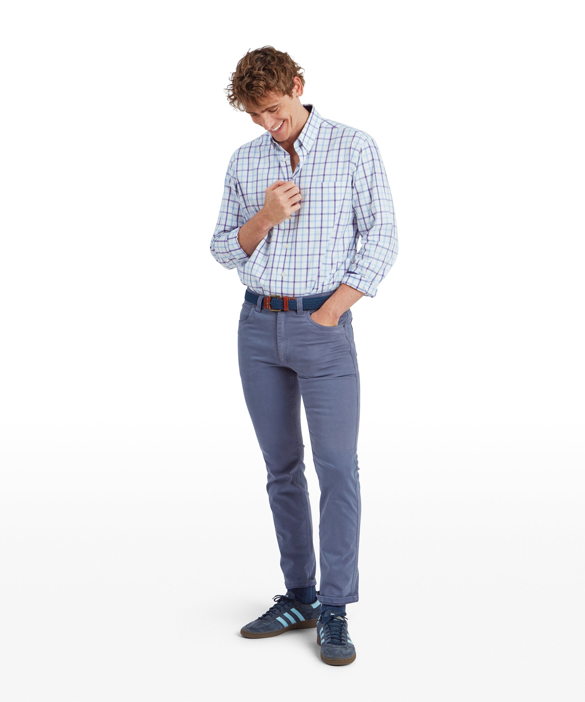 The same young man is standing in a full-body shot, wearing the Schöffel Brancaster Classic Shirt for Men in Purple tucked into slim-fit light blue pants. He is smiling while looking down, holding his shirt lightly with one hand. He is also wearing a blue woven belt with brown leather accents and navy blue trainers, giving a casual yet stylish appearance.