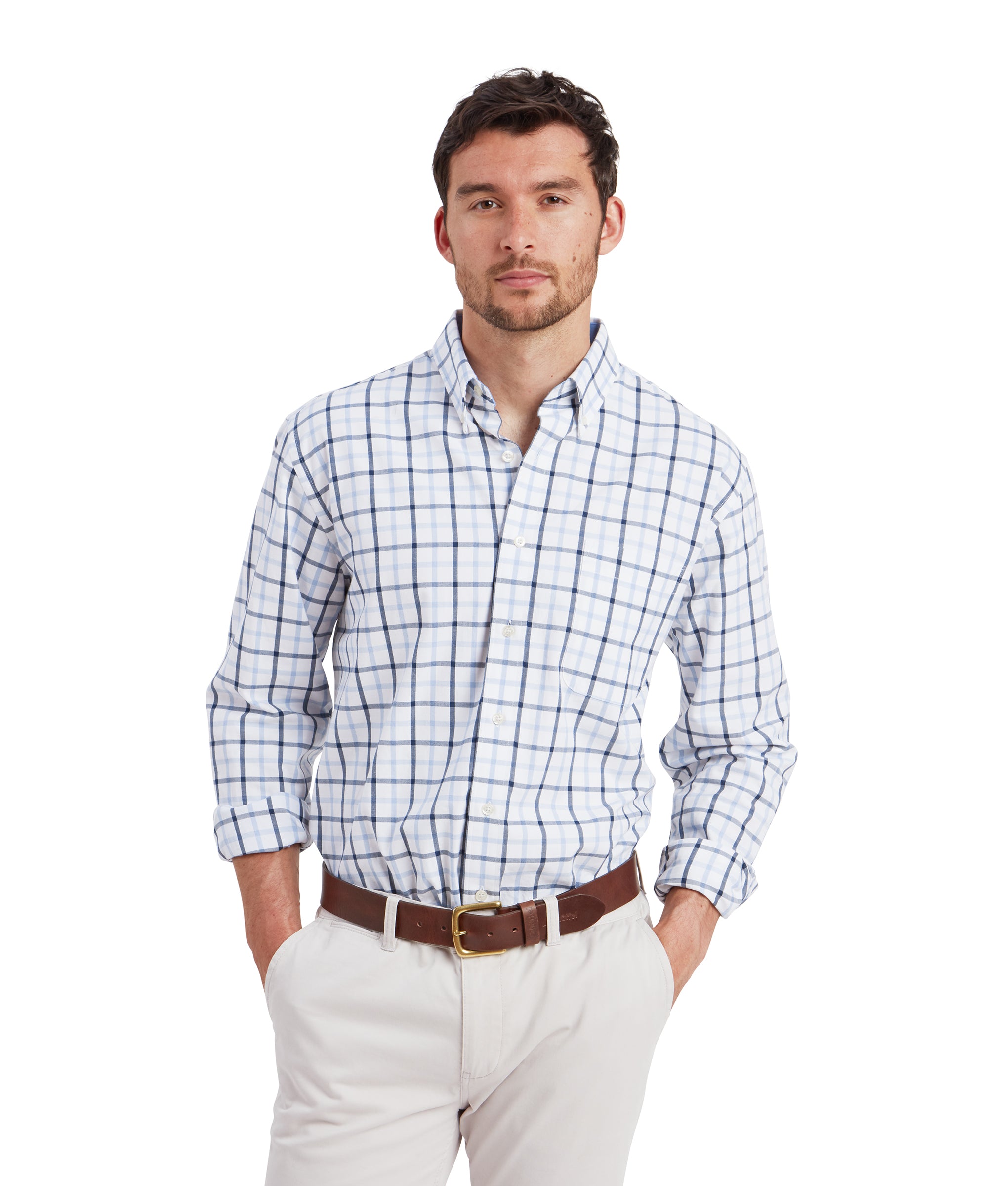 A man stands confidently wearing a Schöffel Brancaster Classic Shirt for Men in Dark Blue with a blue and light gray checkered pattern. The shirt has a classic collar and is tucked into a pair of light beige chinos. He has his hands casually placed in his pockets, and he is also wearing a brown leather belt, adding a polished touch to the outfit.