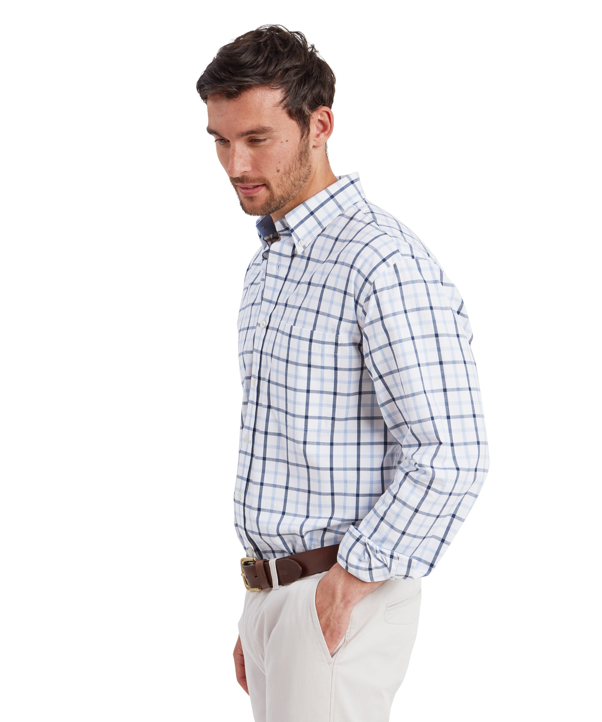 A side profile of the man showcases the Schöffel Brancaster Classic Shirt for Men in Dark Blue’s fit, emphasising the crisp structure of the collar and the neat, tailored fit of the shirt. His left hand rests in his pocket, while the light beige chinos and brown leather belt remain prominent elements of the ensemble.