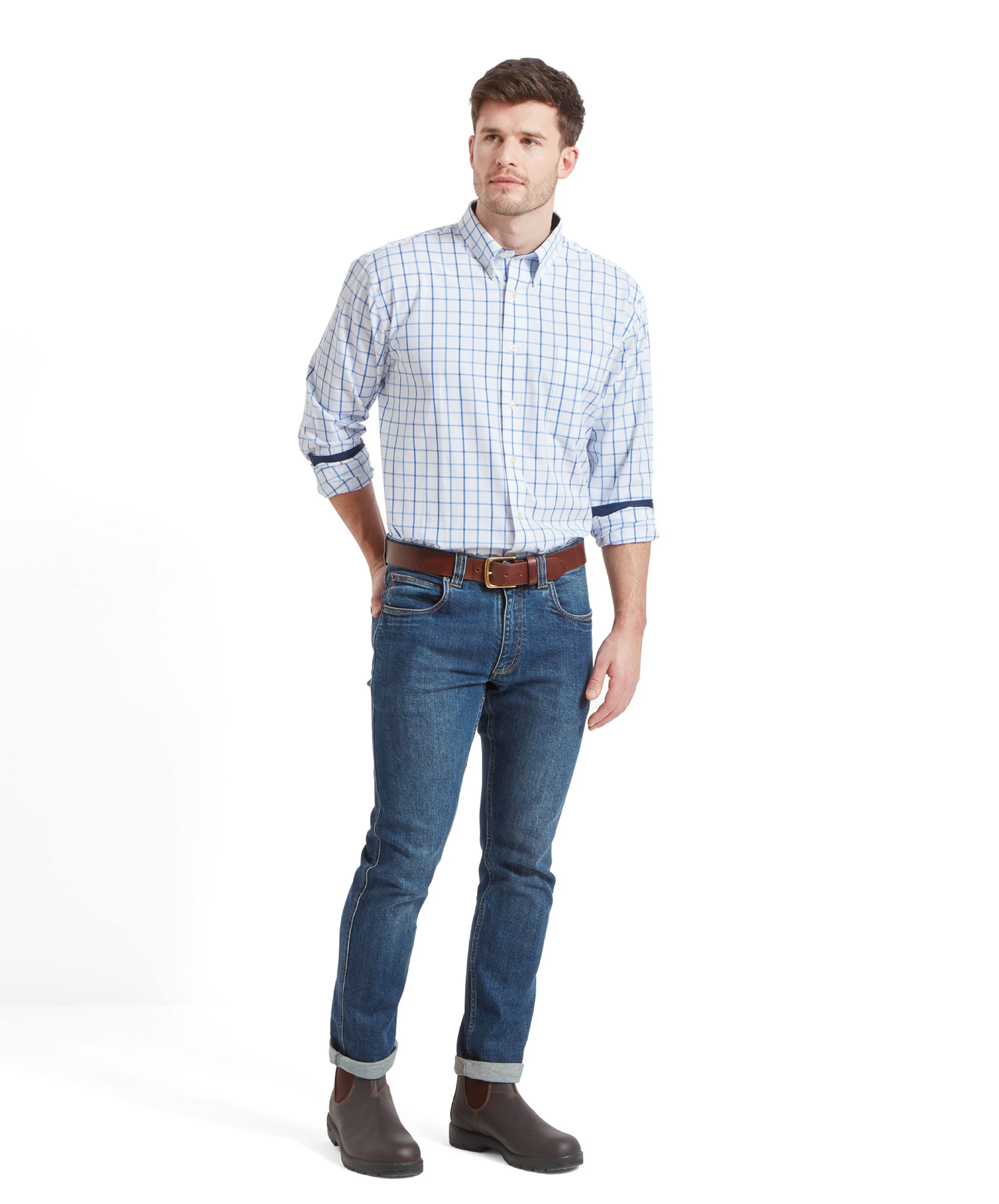 The same man standing in a full-body shot, wearing the same Schöffel Brancaster Classic Shirt for Men in Blue, jeans, and brown belt, with brown boots. His hands are in his pockets, and he is looking slightly to his right.