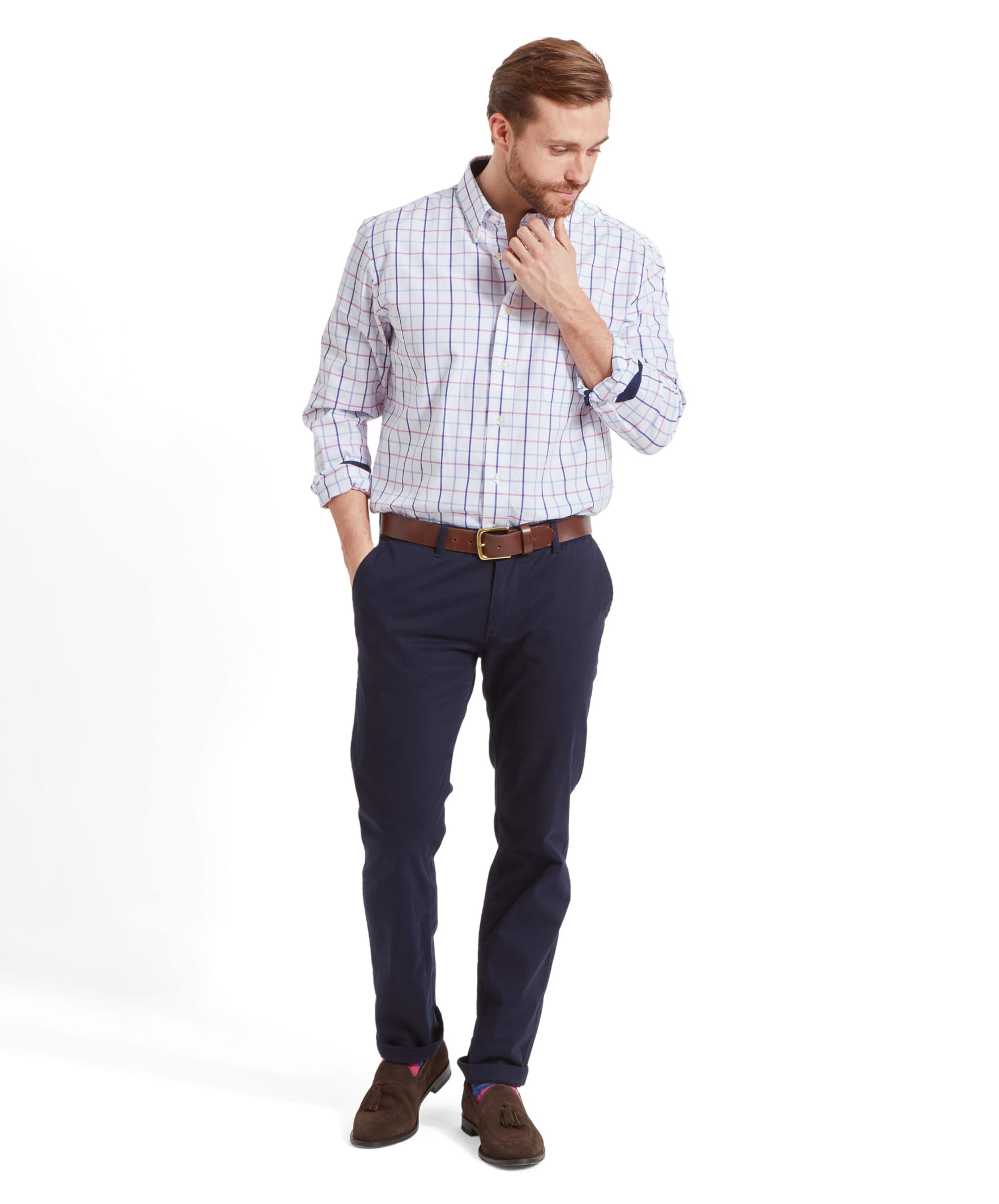 A full-body shot of a man wearing the Schöffel Brancaster Classic Shirt for Men in Pink, paired with dark blue trousers and brown shoes, looking down and touching his chin thoughtfully.