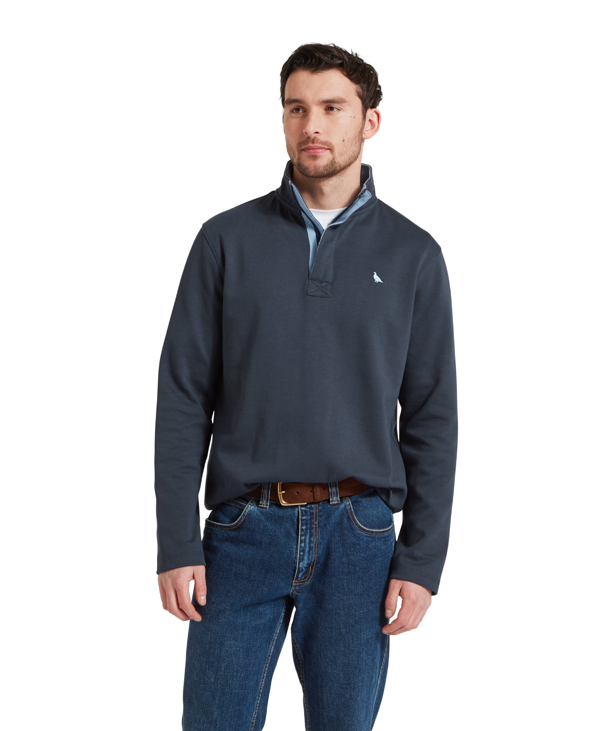 Half height image of a male model wearing a navy Schöffel sweatshirt with white bird logo on the chest. It is paired with a white t-shirt, dark denim jeans and brown leather belt. He is standing in a relaxed pose with his hands by his side looking away from the camera.