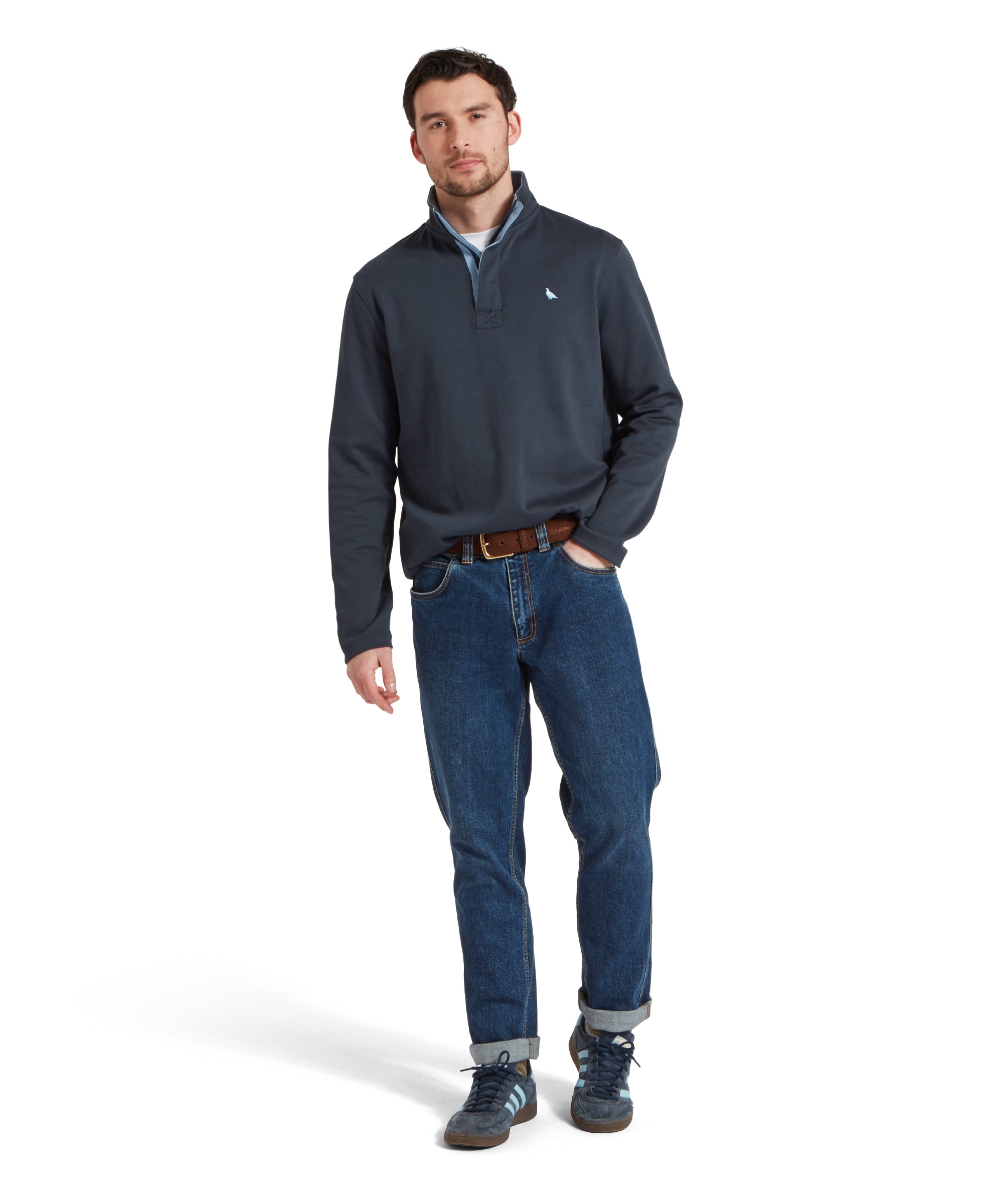 Full height image of a male model wearing a navy Schöffel sweatshirt with white bird logo on the chest. It is paired with a white t-shirt, dark denim jeans, navy trainers and brown leather belt. He is standing in a relaxed pose with one hand in his product looking towards the camera.