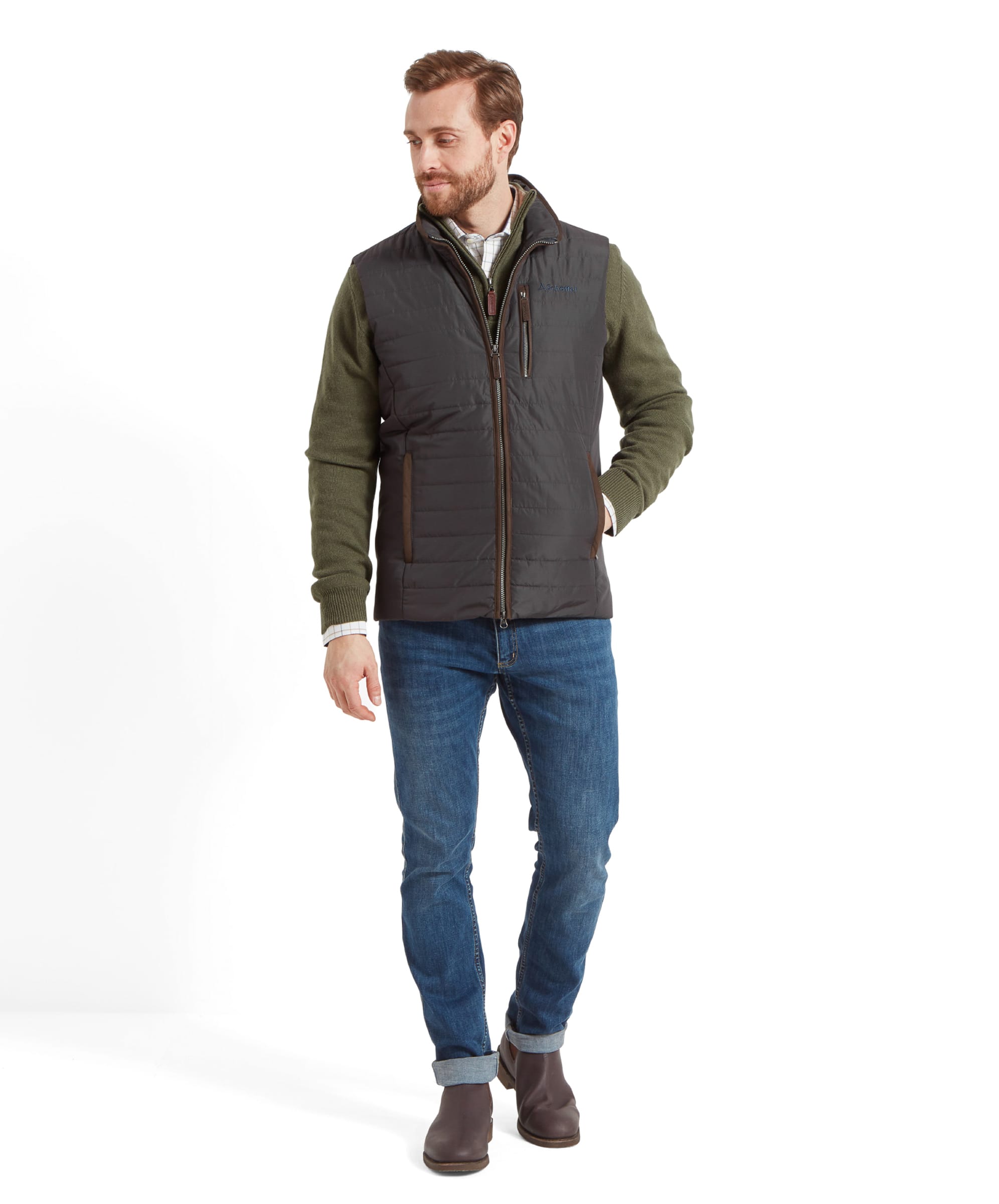 The model displays the full outfit, consisting of the Schöffel Brora Gilet for Men in Grey, green sweater, blue jeans, and brown boots. The gilet has zippered pockets and a sleek, fitted design.