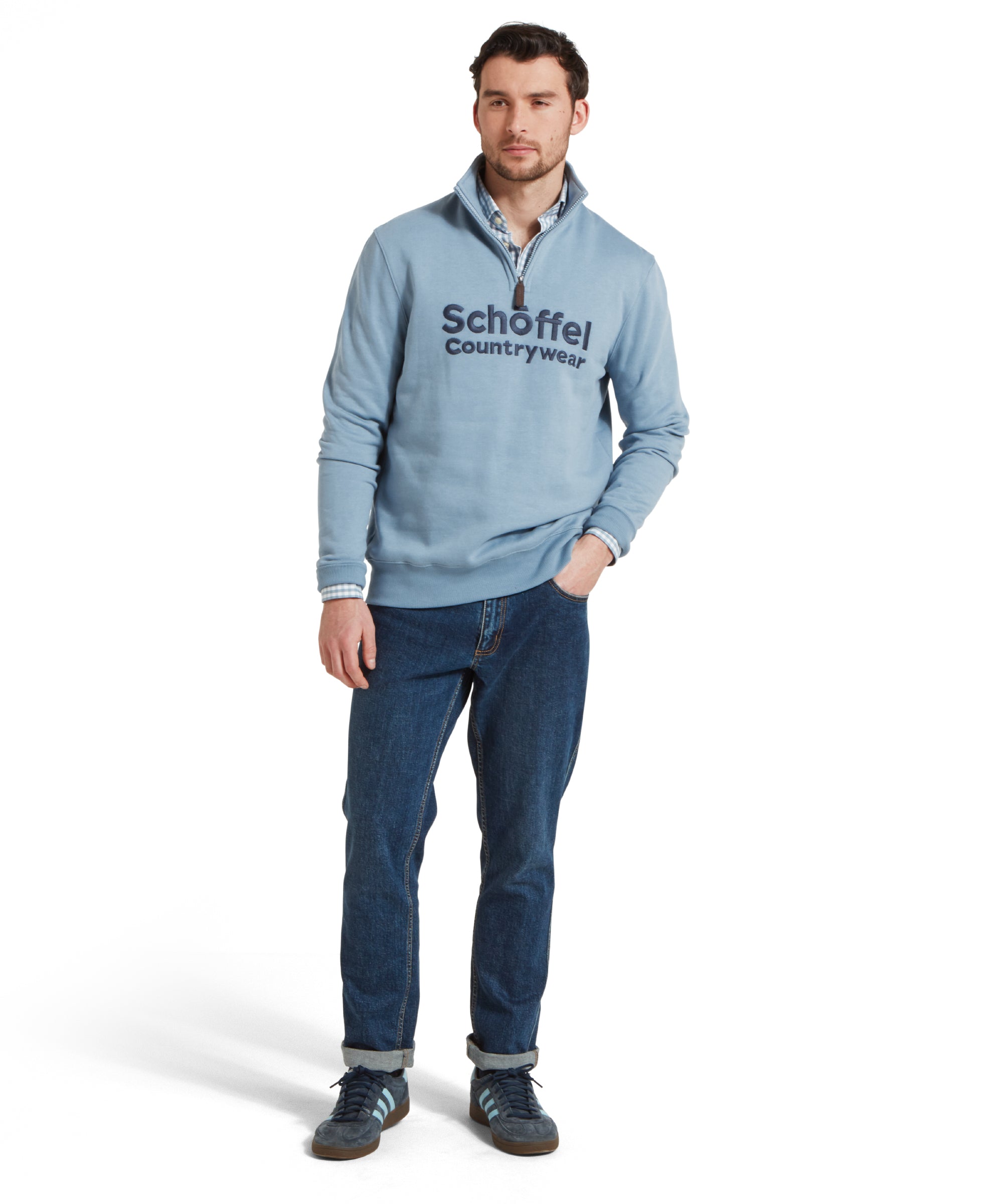 Full height image of a man wearing the Schöffel Bude Sweatshirt for Men in Blue paired with dark blue jeans, navy trainers and checked shirt. He is standing in a relaxed pose with one hand in his pocket, looking slightly to the side.