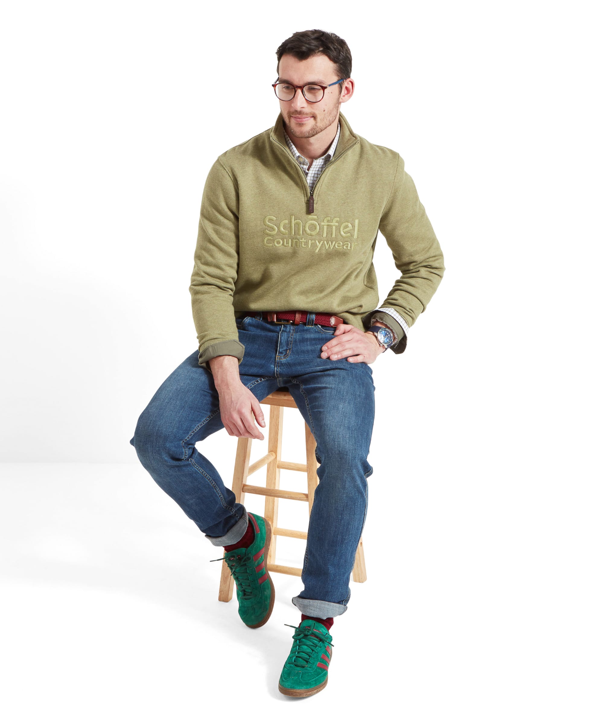 Full-body view of the man sitting on a stool, wearing the Schöffel Bude Sweatshirt for Men in Light Green, paired with jeans and green trainers.