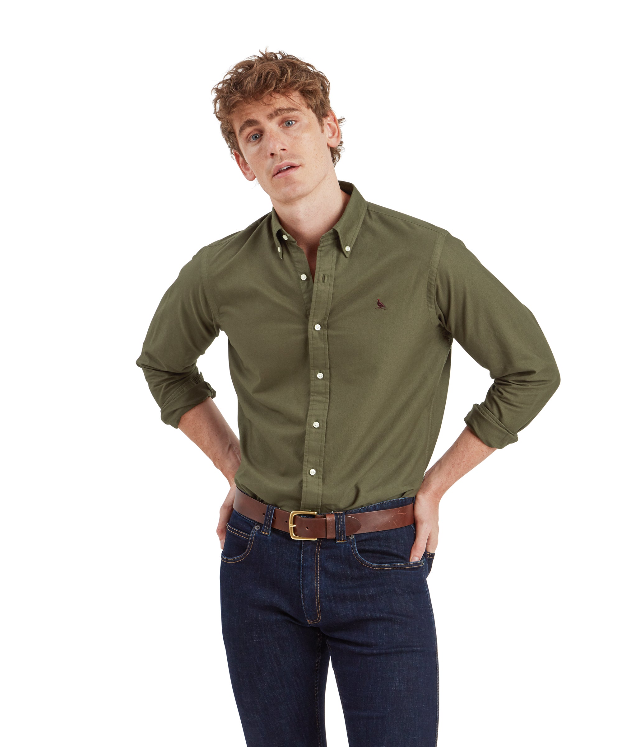 A male model is wearing a fitted long-sleeve Schöffel Cavendish Oxford Shirt for Men in Green. The shirt features a buttoned collar and a small embroidered bird logo on the left chest. He is standing with his hands on his hips, looking straight ahead with a relaxed expression. The shirt is tucked into dark blue jeans, which are paired with a brown leather belt.