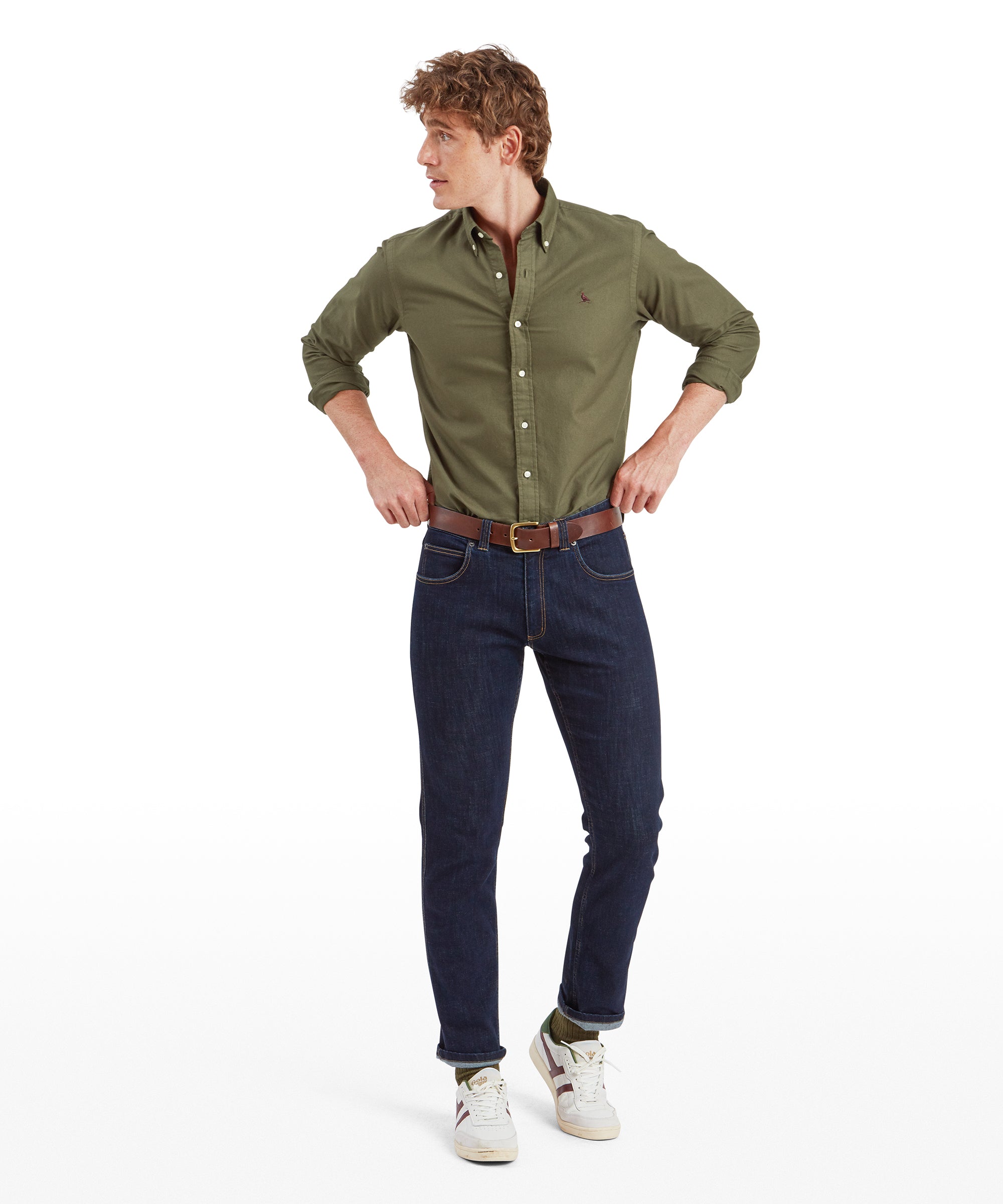 The same male model is now shown in a full-body shot, wearing the Schöffel Cavendish Oxford Shirt for Men in Green tucked into dark blue jeans. He is standing with his legs slightly apart and hands on his hips, looking off to the side. His outfit is completed with a pair of white sneakers with green and brown accents, and a brown leather belt.