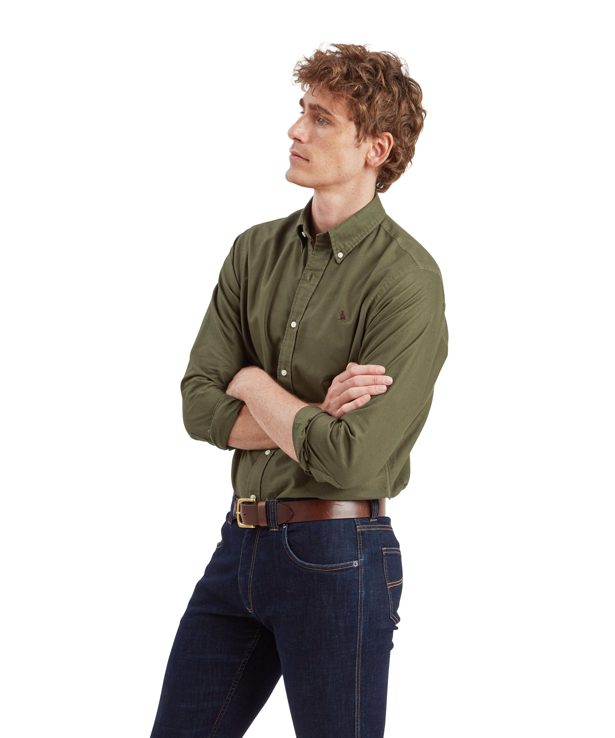 A side view of the male model wearing the Schöffel Cavendish Oxford Shirt for Men in Green, dark blue jeans, and a brown leather belt. He is crossing his arms and looking off to the side, presenting a casual yet thoughtful pose.