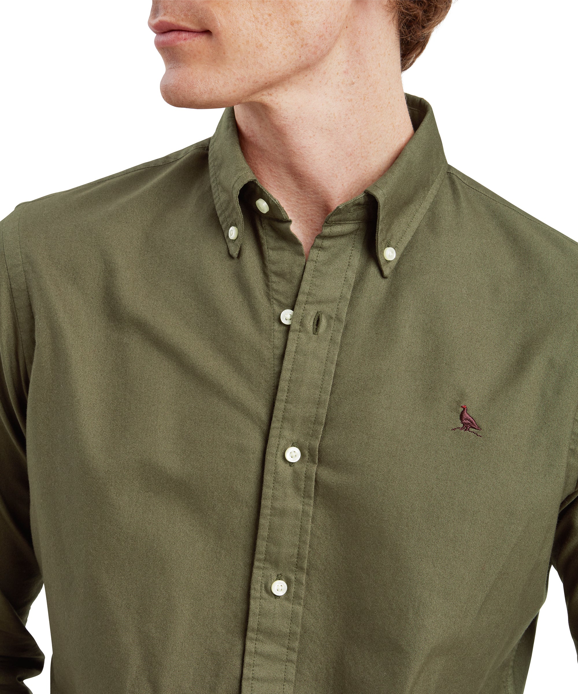 A close-up of the Schöffel Cavendish Oxford Shirt for Men in Green, focusing on the upper chest and collar. The small embroidered bird logo on the left chest is visible, and the model is slightly turning his head to the left. The details of the buttons and the texture of the shirt fabric are clearly shown.