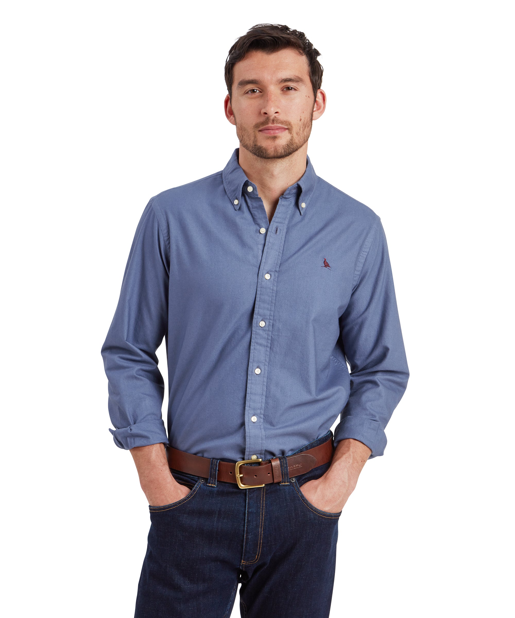 A man is wearing a Schöffel Cavendish Oxford Shirt for Men in Navy with a buttoned collar. The shirt features a small red embroidered bird on the left chest. He has his hands in his jean pockets and is wearing a brown leather belt. He looks directly at the camera with a neutral expression.