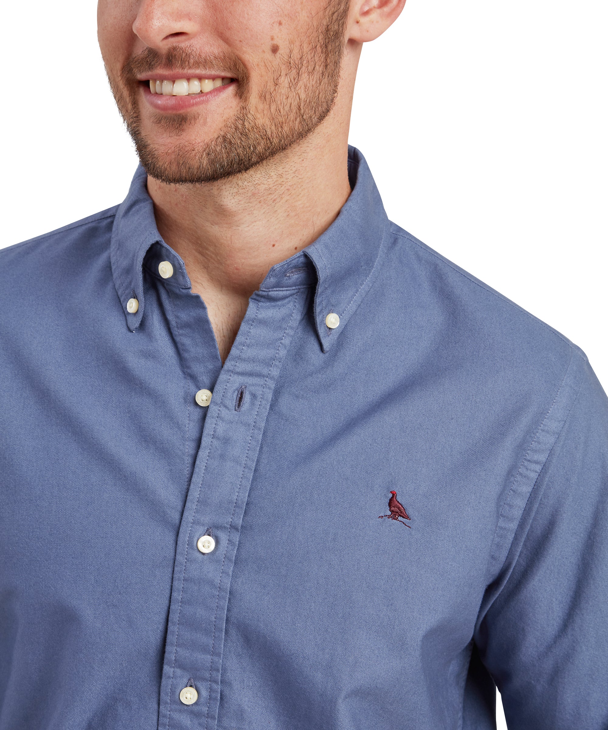 A close-up shot of the man’s upper torso and face. He smiles slightly, focusing on the Schöffel Cavendish Oxford Shirt for Men in Navy’s details, such as the buttoned collar and the embroidered bird on the chest. The image highlights the texture of the fabric and the precision of the shirt's stitching.