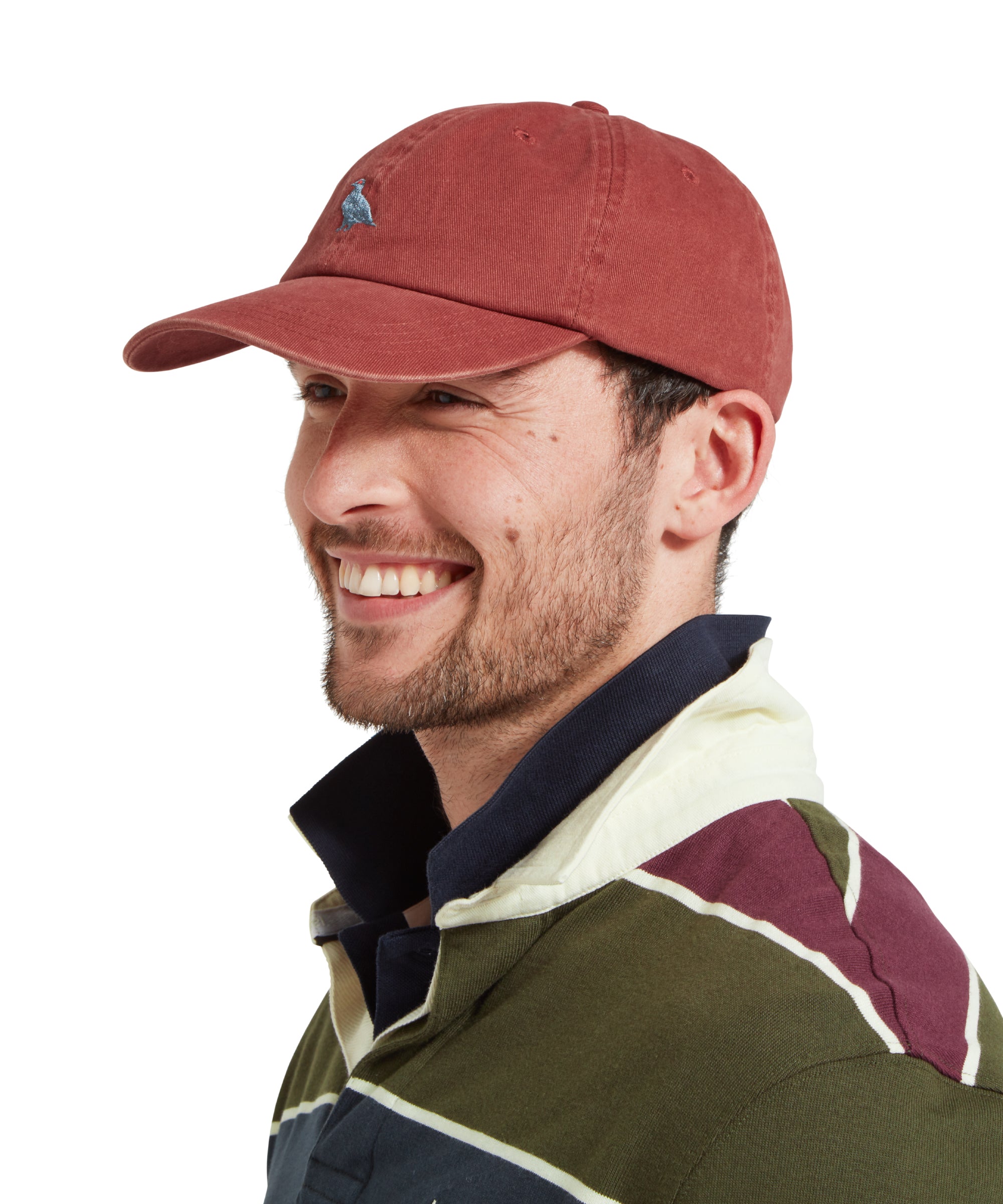 A side profile of a male model wearing a red cap with blue embroidered bird logo on the front. It is paired with a green, blue and burgundy stiped rugby shirt and blue polo shirt. He is looking away from the camera and smiling.