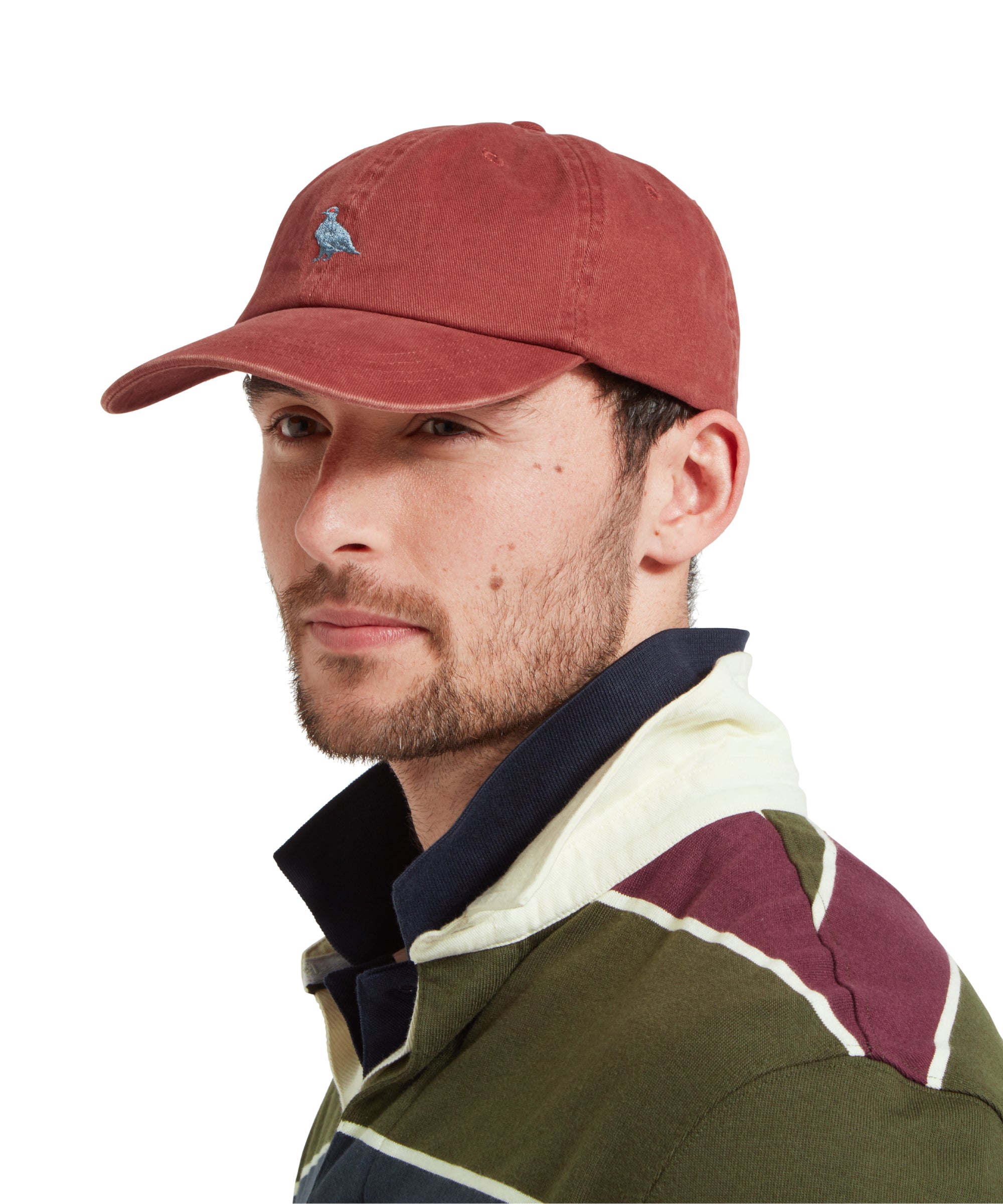 A side profile of a male model wearing a red cap with blue embroidered bird logo on the front. It is paired with a green, blue and burgundy stiped rugby shirt and blue polo shirt.