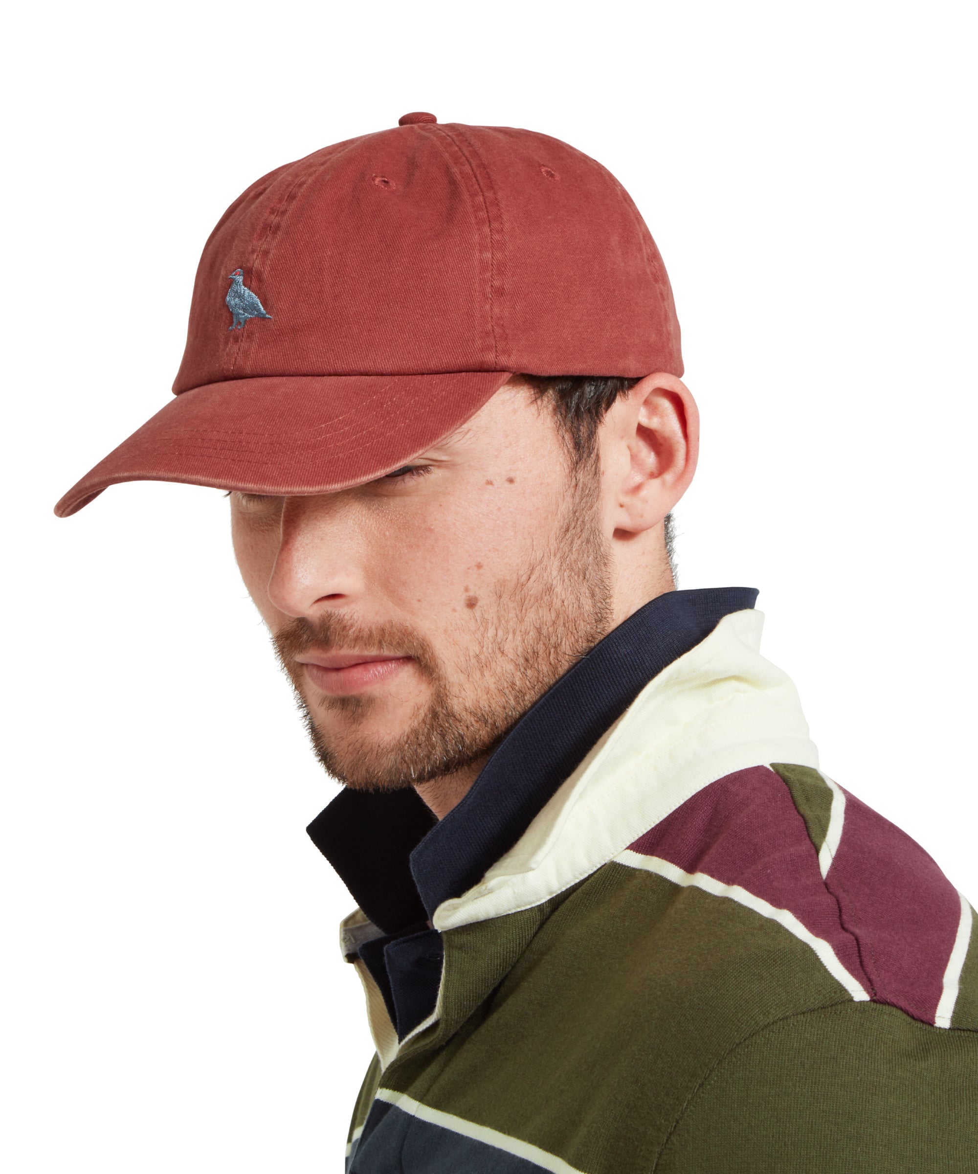 A side profile of a male model wearing a red cap with blue embroidered bird logo on the front. It is paired with a green, blue and burgundy stiped rugby shirt and blue polo shirt.