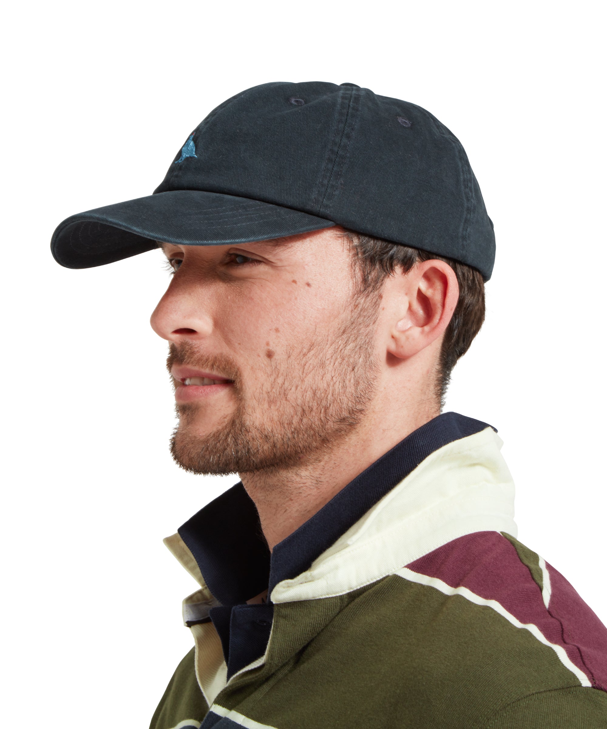 A side profile of a male model wearing a navy cap with a light blue embroidered bird logo on the front. It is paired with a green, blue and burgundy stiped rugby shirt and blue polo shirt.