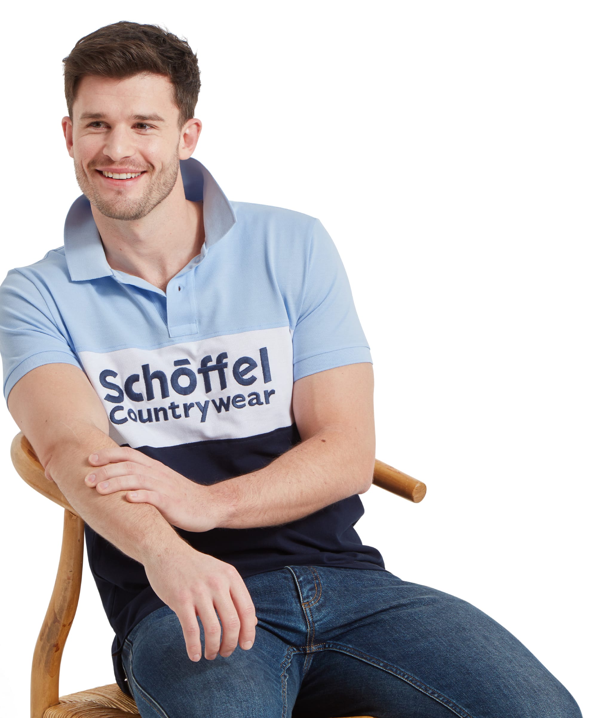 A man smiling and sitting on a chair, wearing a Schöffel Men's Exeter Heritage Polo Shirt in Blue with "Schöffel Countrywear" written across the chest.