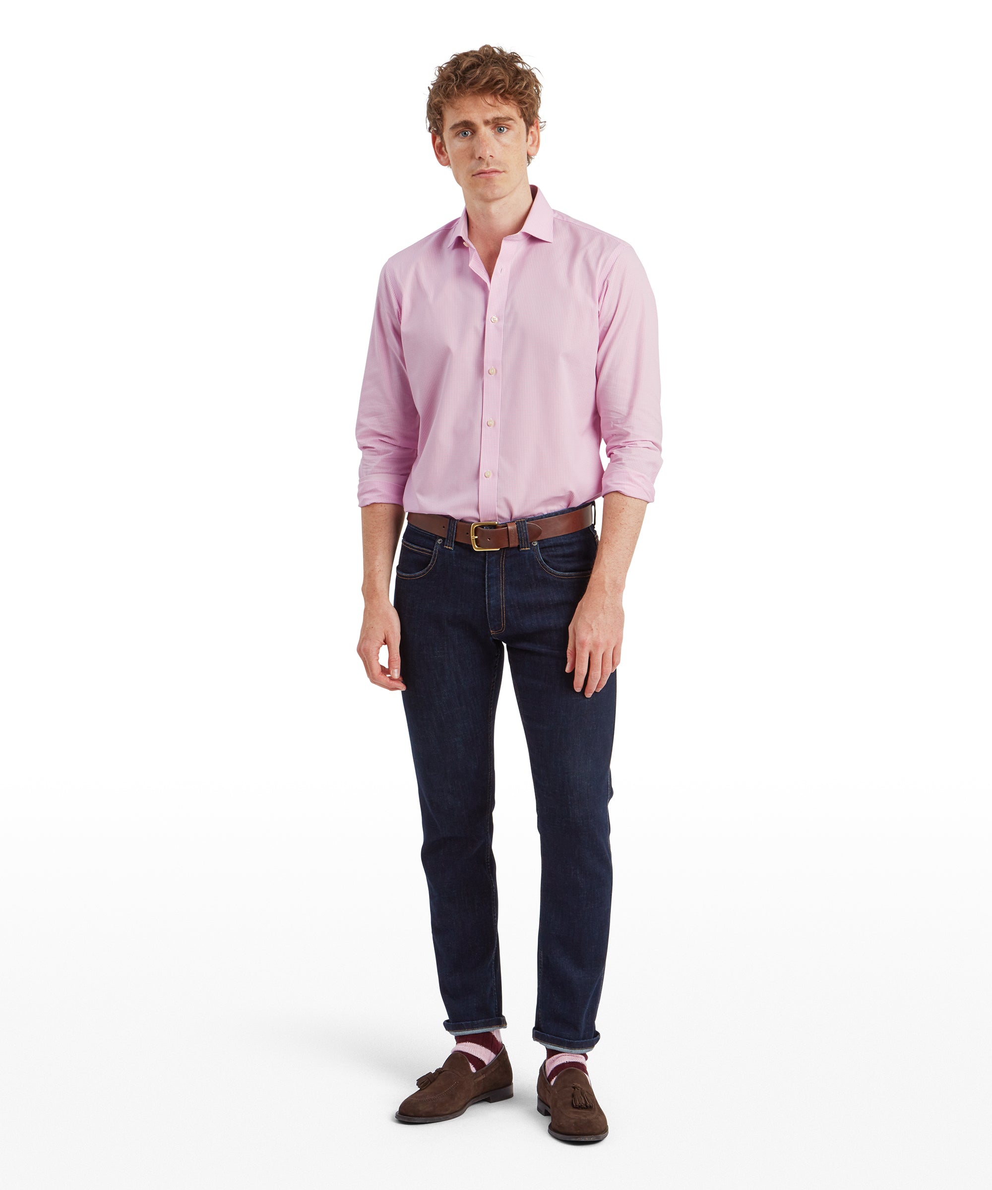 A full-body shot of the man wearing the Schöffel Greenwich Tailored Shirt for Men in Pink Check. He stands facing forward with a neutral expression, his hands relaxed by his sides. The shirt is tucked into his dark denim jeans, which are slim-fitting and cuffed at the ankles. He completes the look with brown suede loafers, pink striped socks, and a brown leather belt.