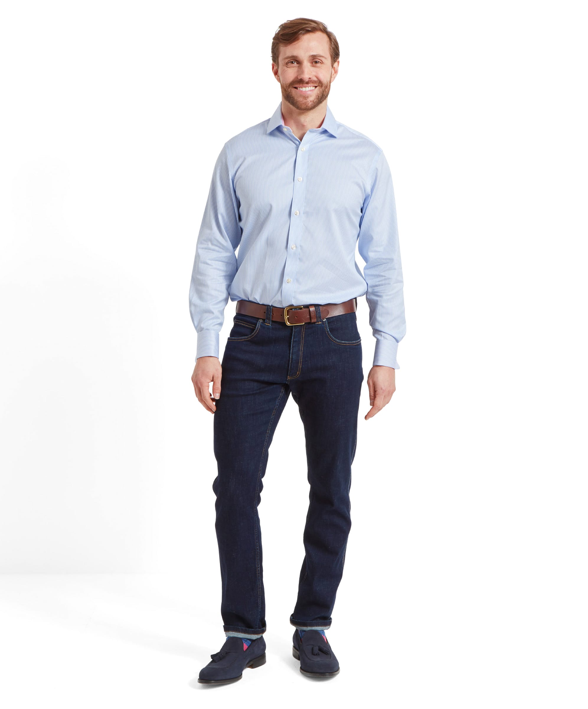 A full-body view of a man wearing a Schöffel Greenwich Tailored Shirt for Men in Blue Stripe, standing and smiling with his hands relaxed by his sides. He is also wearing dark blue jeans, a brown belt, and navy loafers.