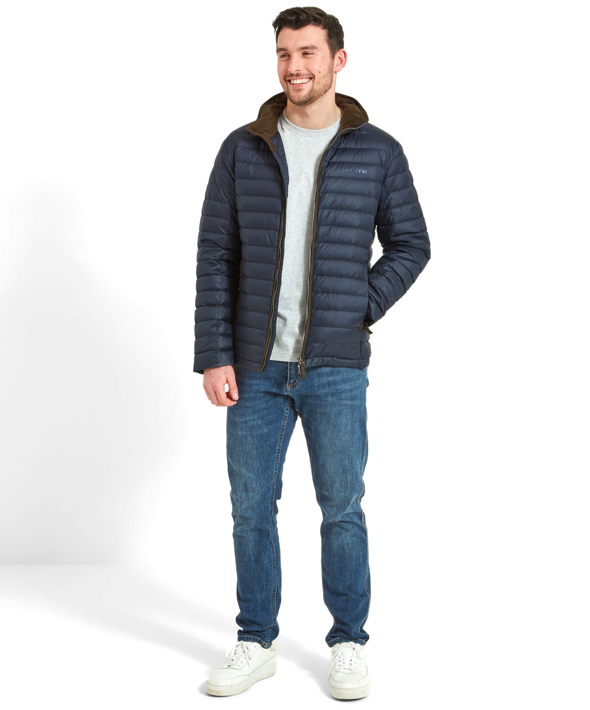 Full-body view of a man wearing the Schöffel Hallaton Down Jacket for Men in Blue with blue jeans,. White trainers and a grey t-shirt. The man is smiling looking to his left with his right hand in the jacket pocket.