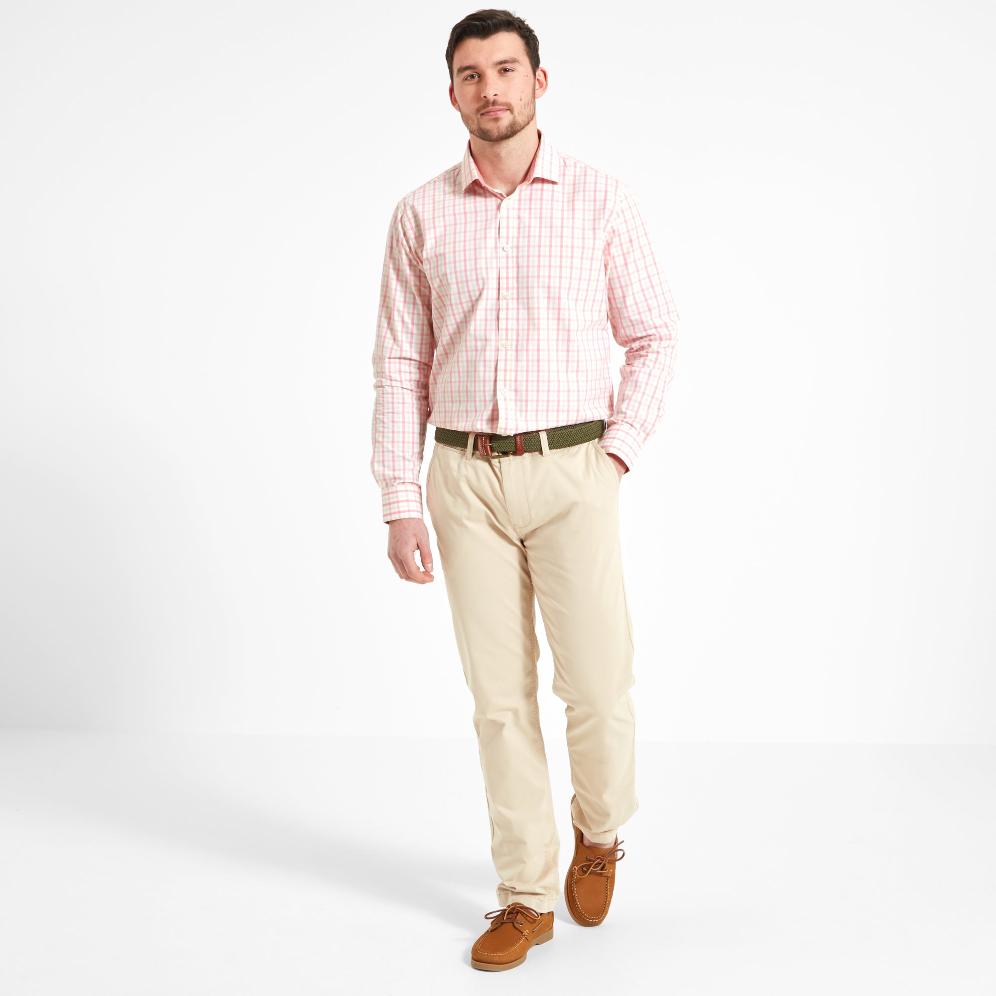 Full body view of a man wearing the same Schöffel Hebden Tailored Shirt for Men in Pink Check with beige trousers, a green wovne belt and tan shoes. He has his right hand in the trouser pocket and is walking forward.