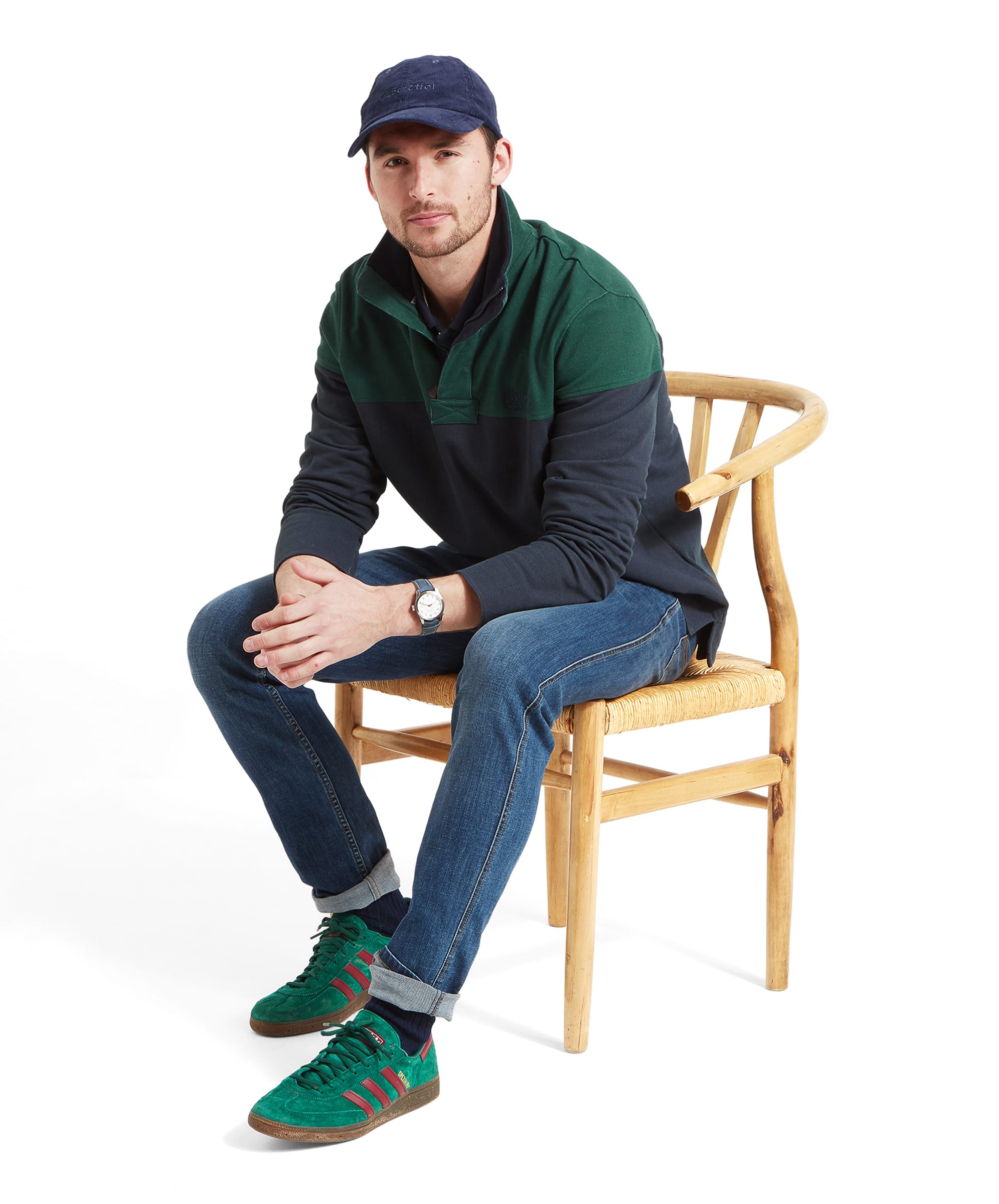 A man sits of a wooden chair with his elbows on his knees whilst wearing the Schöffel Helford Heritage Sweatshirt for Men in Green with a blue cap, navy jeans and green trainers.